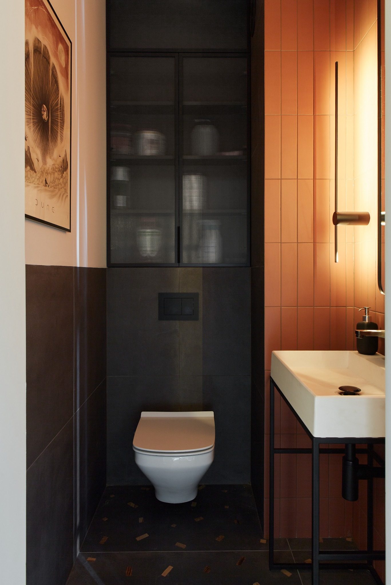 Nowoczesna łazienka z wiszącą toaletą, umywalką na metalowym stelażu i pomarańczowymi płytkami, oświetlona lampą ścienną, z plakatem filmowym na ścianie.