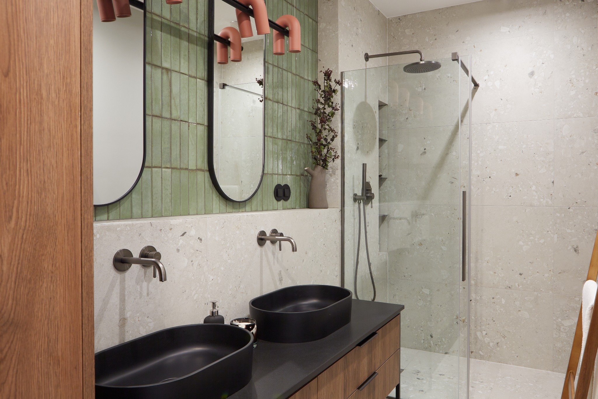Nowoczesna łazienka z dwoma czarnymi umywalkami nablatowymi, bateriami w kolorze grafitu, zielonymi płytkami ściennymi ułożonymi pionowo, dwoma owalnymi lustrami z czarnymi ramami i kabiną...