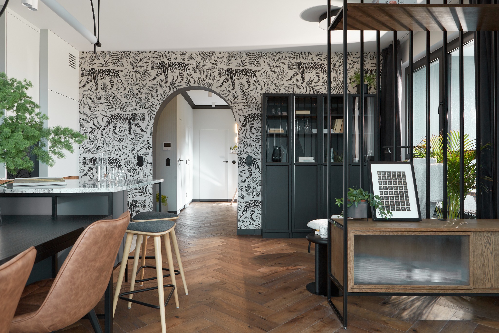 Nowoczesne wnętrze z parkietem w jodełkę, tapetą w motywy zwierzęce i roślinne, czarną witryną i drewnianą komodą z ryflowanym szkłem, oddzielone metalową konstrukcją od jasnego pomieszczenia...