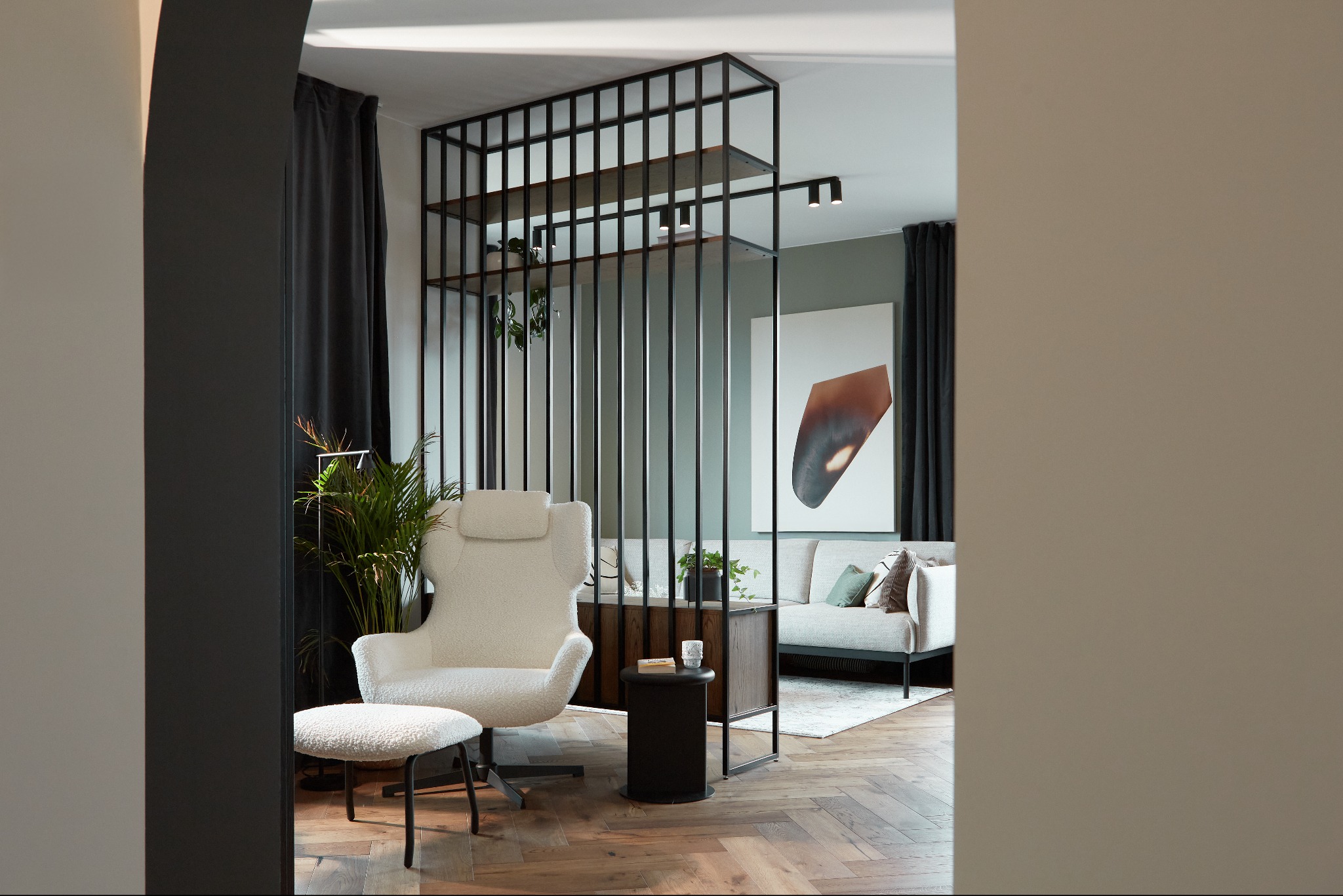 Nowoczesny salon z drewnianą podłogą w jodełkę, dzielony ażurowym regałem z czarnymi metalowymi prętami i drewnianymi półkami, z białym fotelem typu uszak i podnóżkiem, oraz szarą sofą w tle.