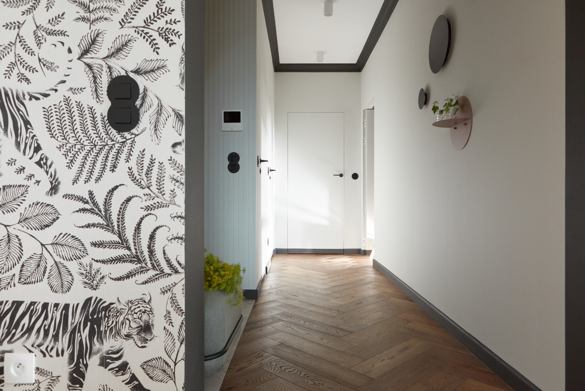 Nowoczesny korytarz z parkietem w jodełkę, ściana z tapetą w motywy roślinne i tygrysa, białe drzwi z czarnymi klamkami, dekoracyjne półki i czarne okrągłe ozdoby na ścianie.