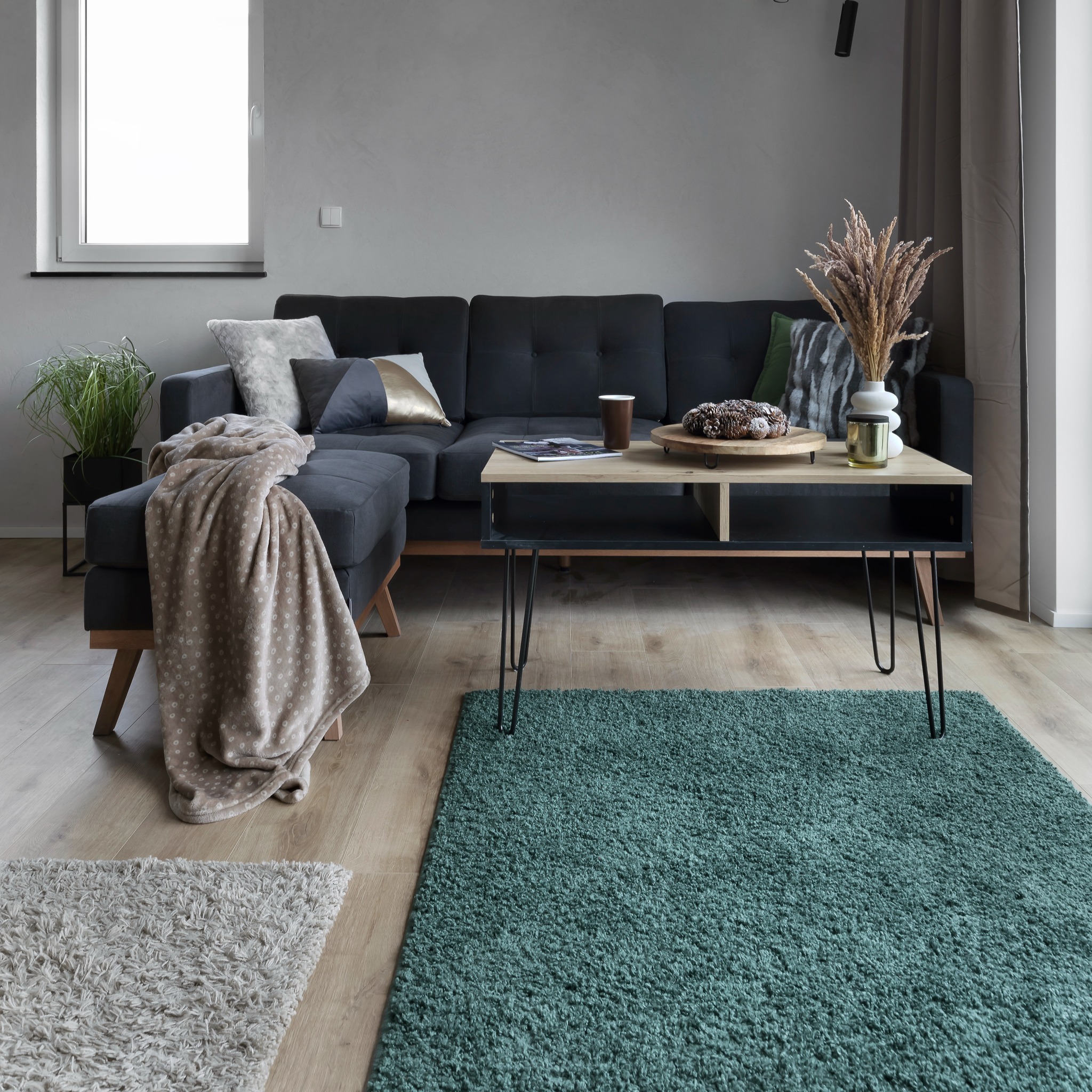 Salon w Gdyni z szarą sofą narożną, drewnianym stolikiem kawowym na metalowych nogach, zielonym dywanem typu shaggy i beżowym pledem z drobnymi kropkami.