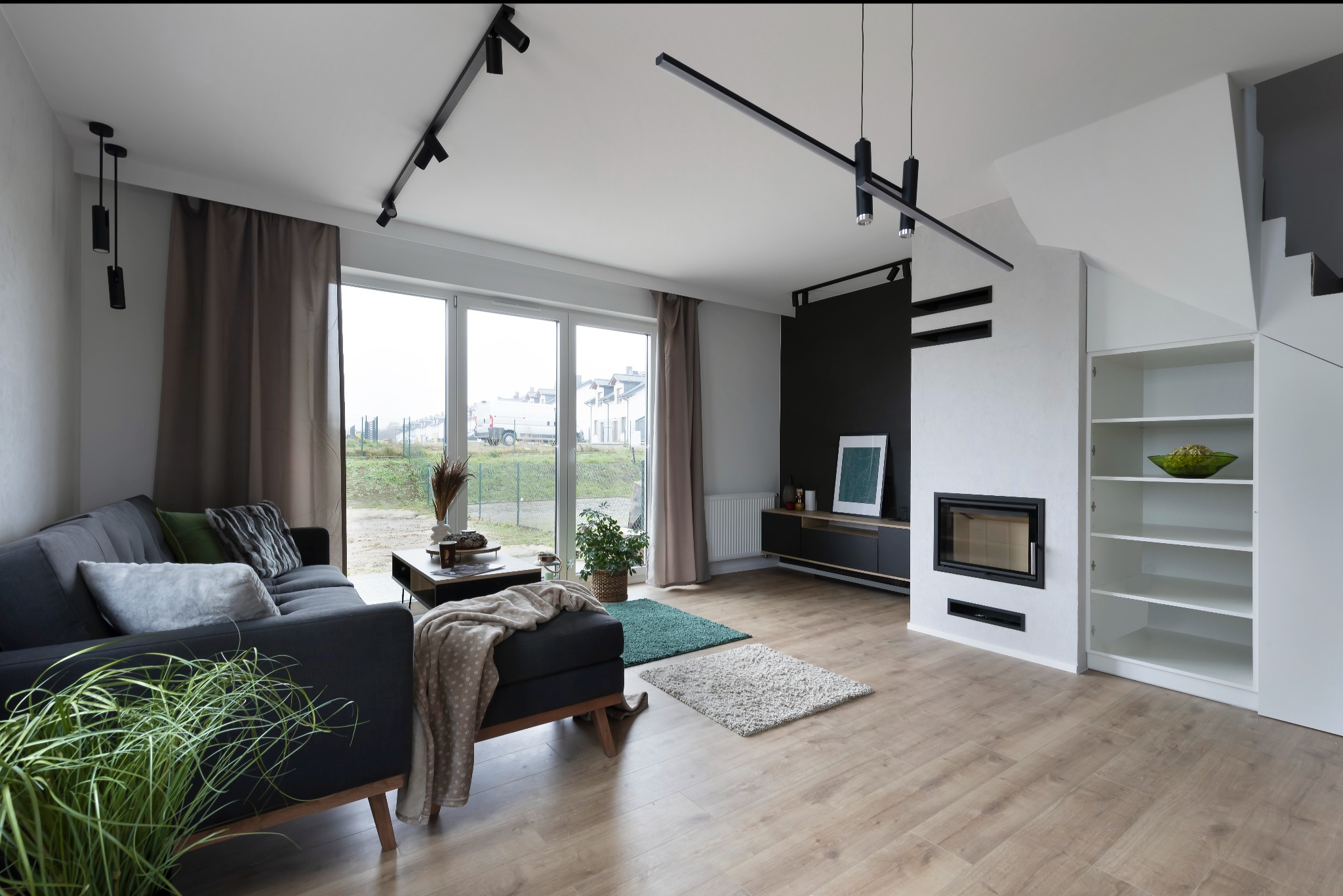 Jasne, przestronne wnętrze salonu z drewnianą podłogą, szarą sofą z poduszkami, nowoczesnym oświetleniem szynowym na suficie i kominkiem obudowanym białą zabudową z półkami.