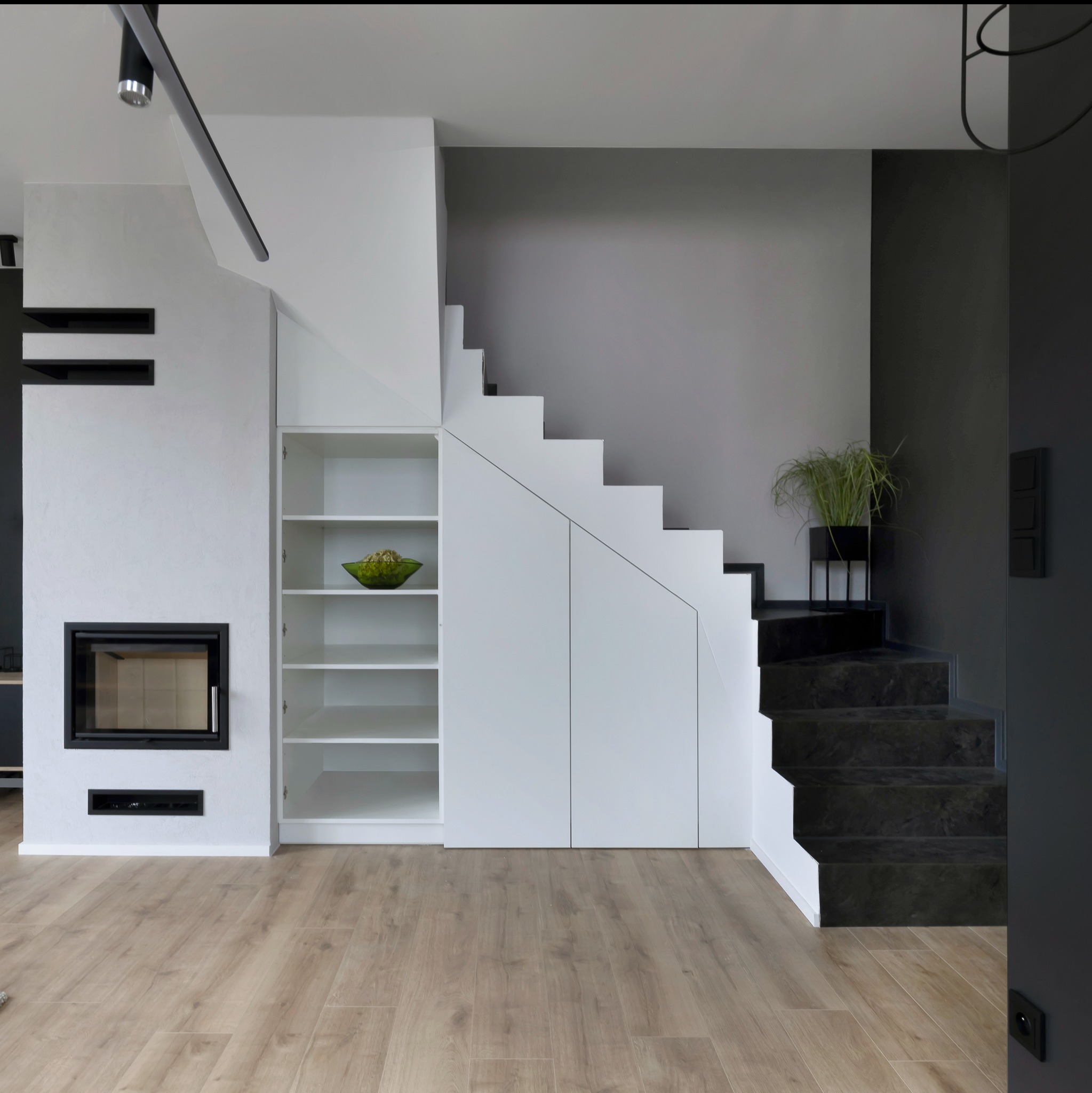 Nowoczesne wnętrze: białe ściany z fakturą, kominek z czarną obudową, drewniana podłoga, schody z ciemnego kamienia i zabudowana szafa pod schodami z otwartymi półkami, na których stoi zielona misa...