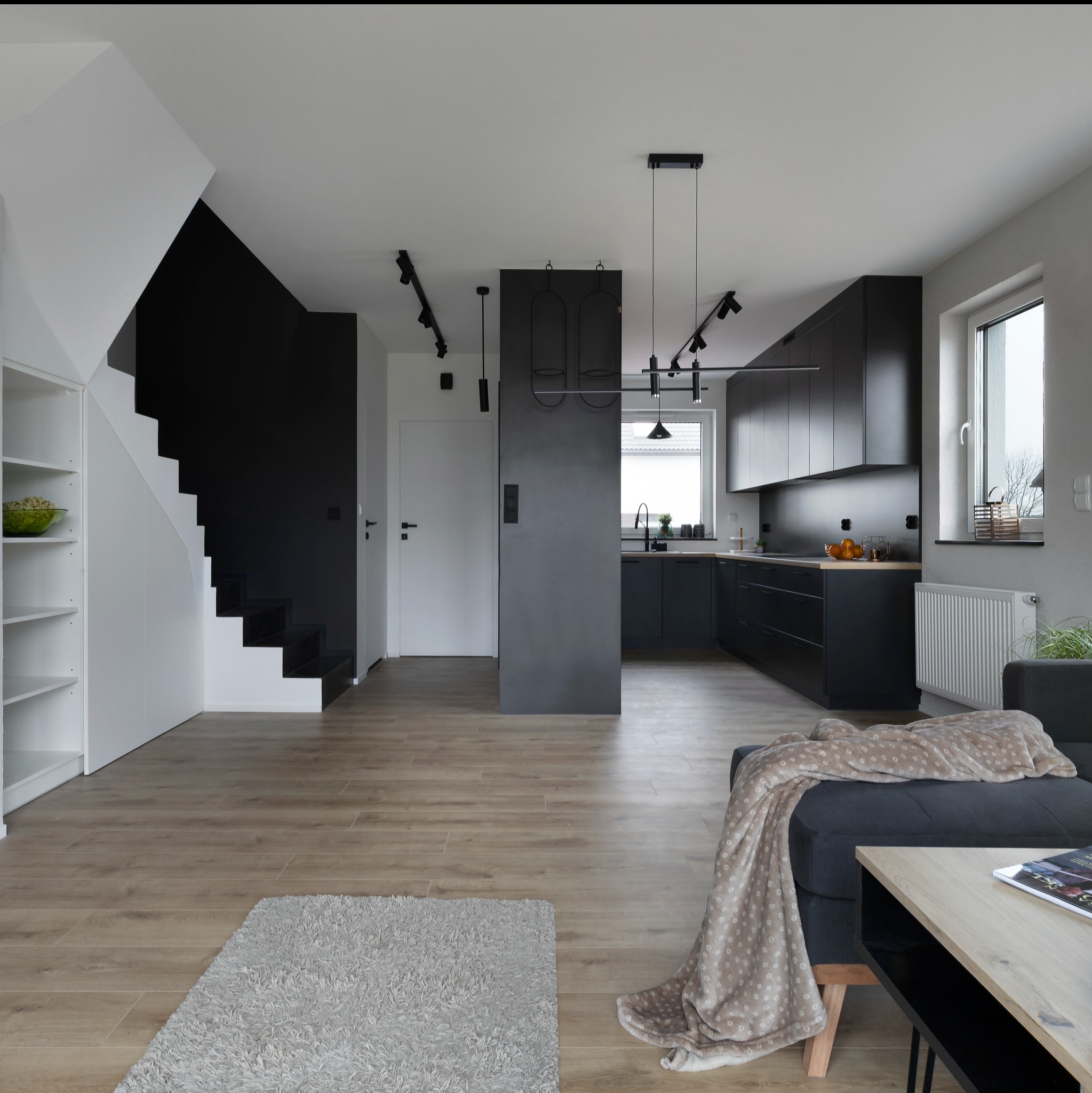 Nowoczesne wnętrze domu: czarna kuchnia z drewnianym blatem, minimalistyczne oświetlenie, schody w czerni i bieli, jasna podłoga z drewnopodobnych paneli, szary dywan i sofa z beżowym pledem.