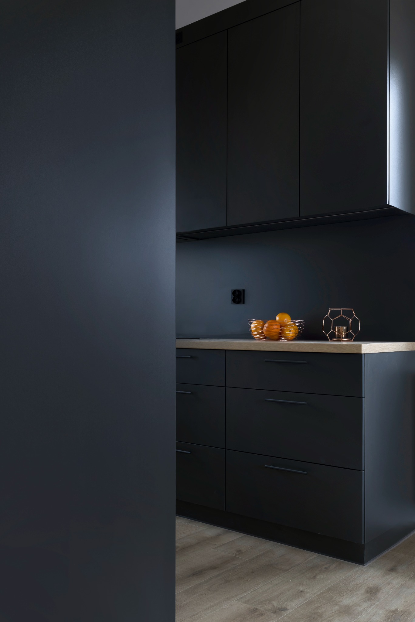 Ciemnogranatowa kuchnia z drewnianym blatem, na którym stoi misa z pomarańczami i geometryczny świecznik, widok z boku na ścianę w tym samym kolorze.