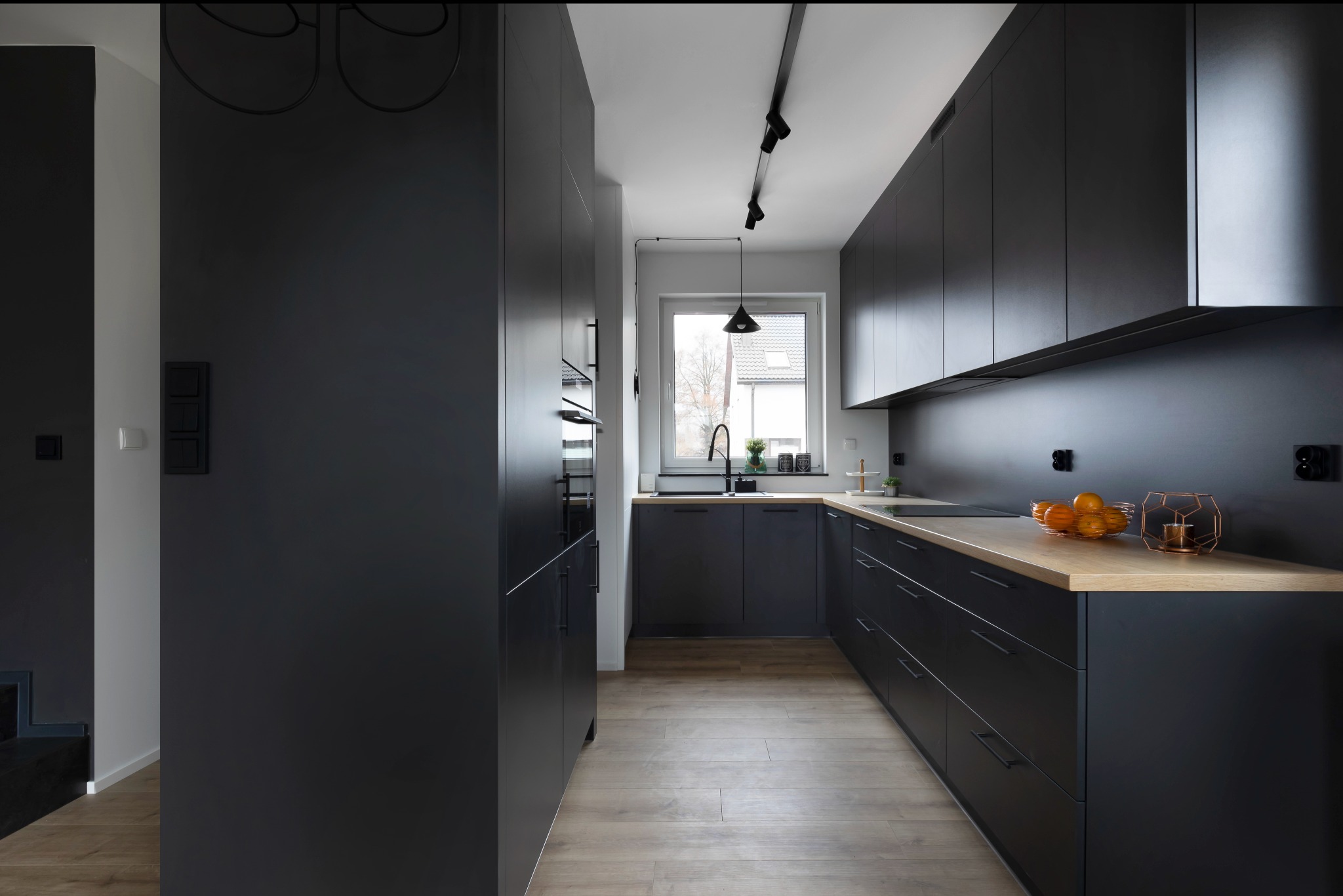 Nowoczesna kuchnia w odcieniach szarości i czerni z drewnianym blatem, oświetleniem punktowym na szynoprzewodzie i widokiem z okna na zabudowę jednorodzinną.