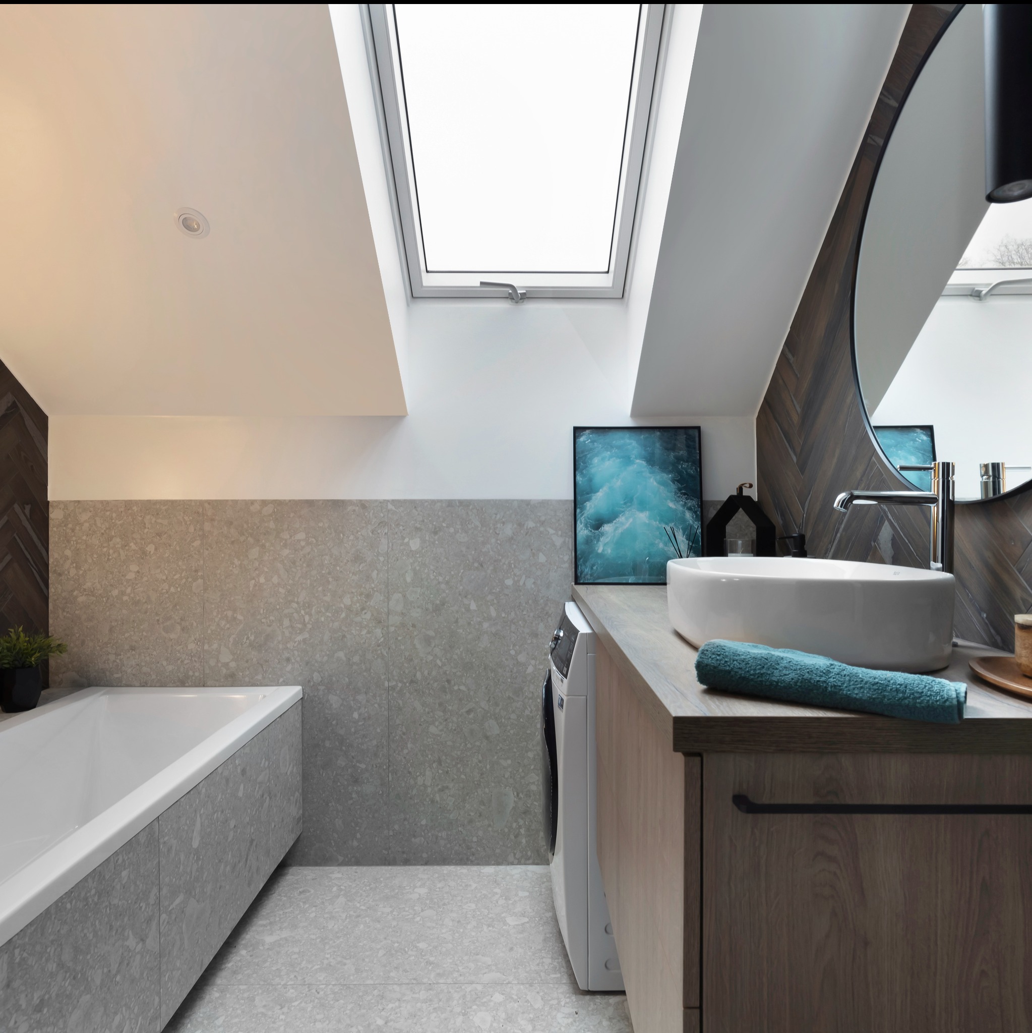 Nowoczesna łazienka na poddaszu z wanną obudowaną płytkami lastryko, umywalką nablatową, drewnianym blatem i szafką, pralką oraz oknem dachowym.