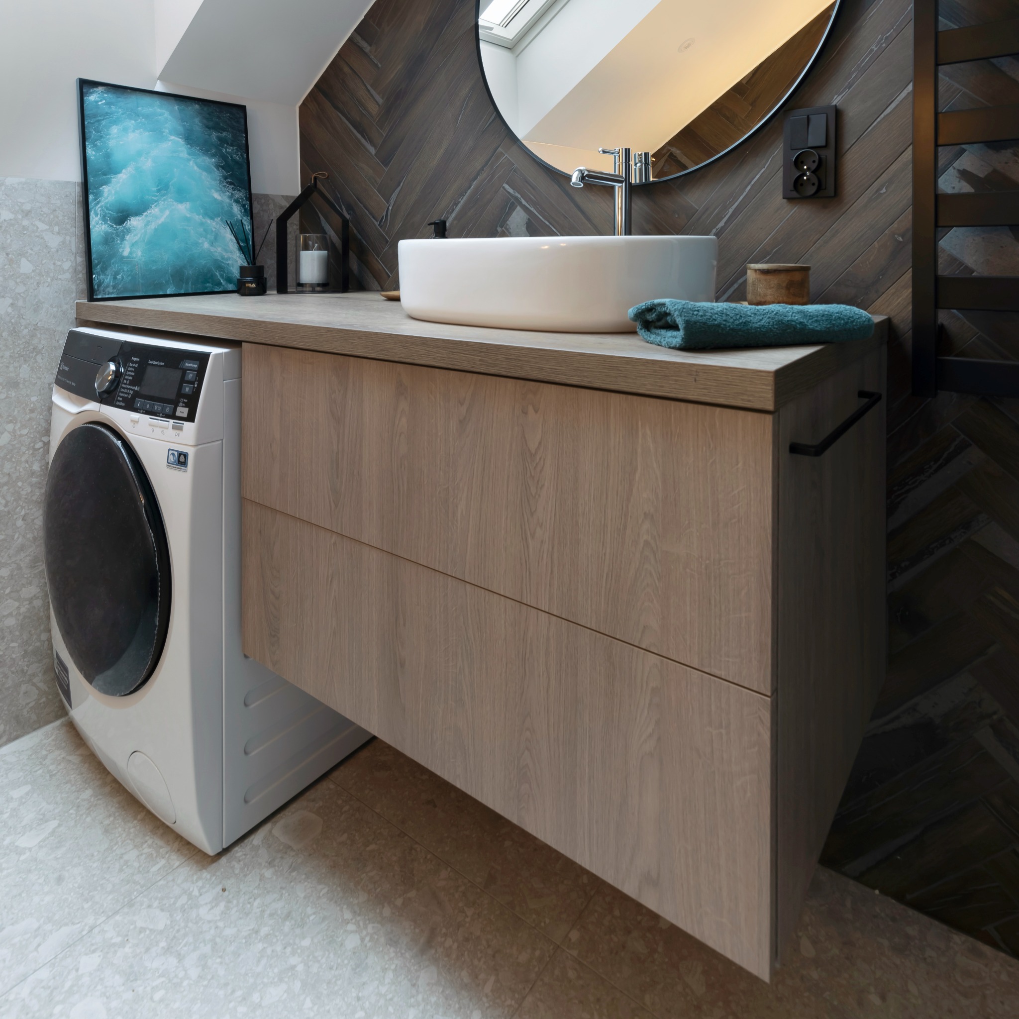 Nowoczesna łazienka z umywalką nablatową, drewnianą szafką z szufladami i pralką obok, na ścianie płytki ułożone w jodełkę, okrągłe lustro z podświetleniem i obraz z motywem morskim.