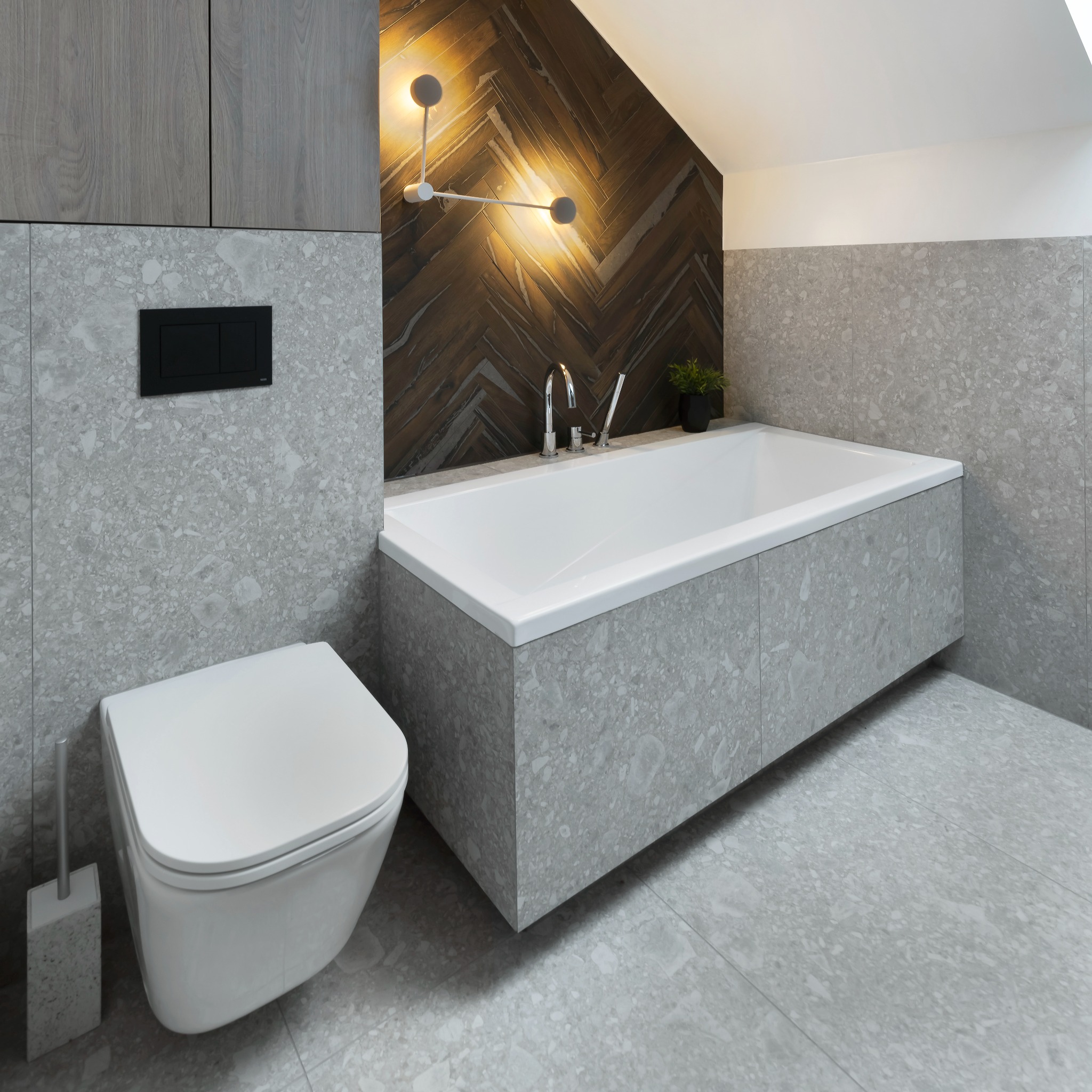 Nowoczesna łazienka w szarościach z podwieszaną toaletą, wanną obudowaną płytkami i dekoracyjną ścianą z drewnopodobnych paneli ułożonych w jodełkę, oświetlona designerskimi lampami.