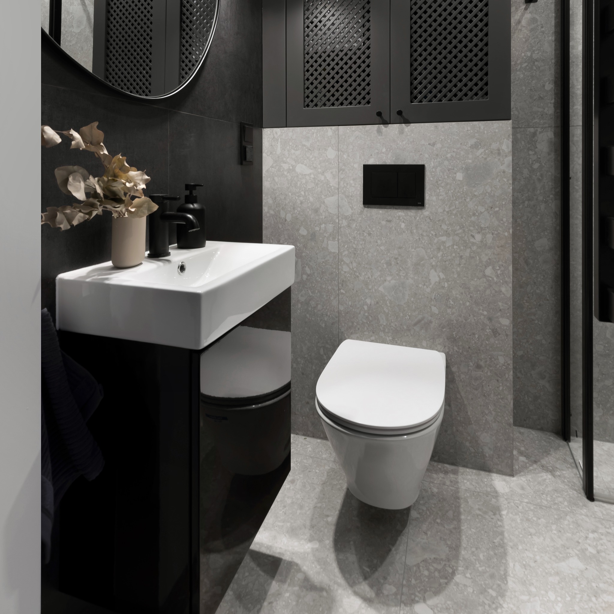 Nowoczesna łazienka w odcieniach szarości i czerni z wiszącą białą miską WC, umywalką na czarnym postumencie, czarną armaturą i szarą płytką imitującą lastryko na ścianach i podłodze, uzupełniona...