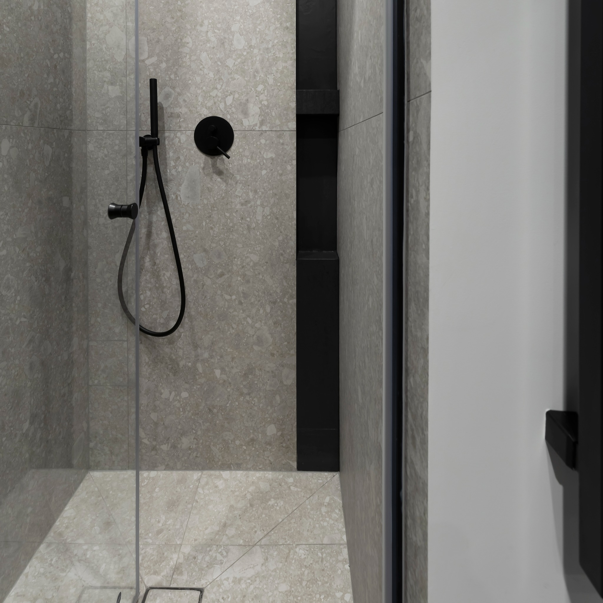 Wnętrze kabiny prysznicowej z szarymi płytkami lastryko na ścianach i podłodze, czarna armatura oraz wnęka w ścianie wykończona czarnymi płytkami.