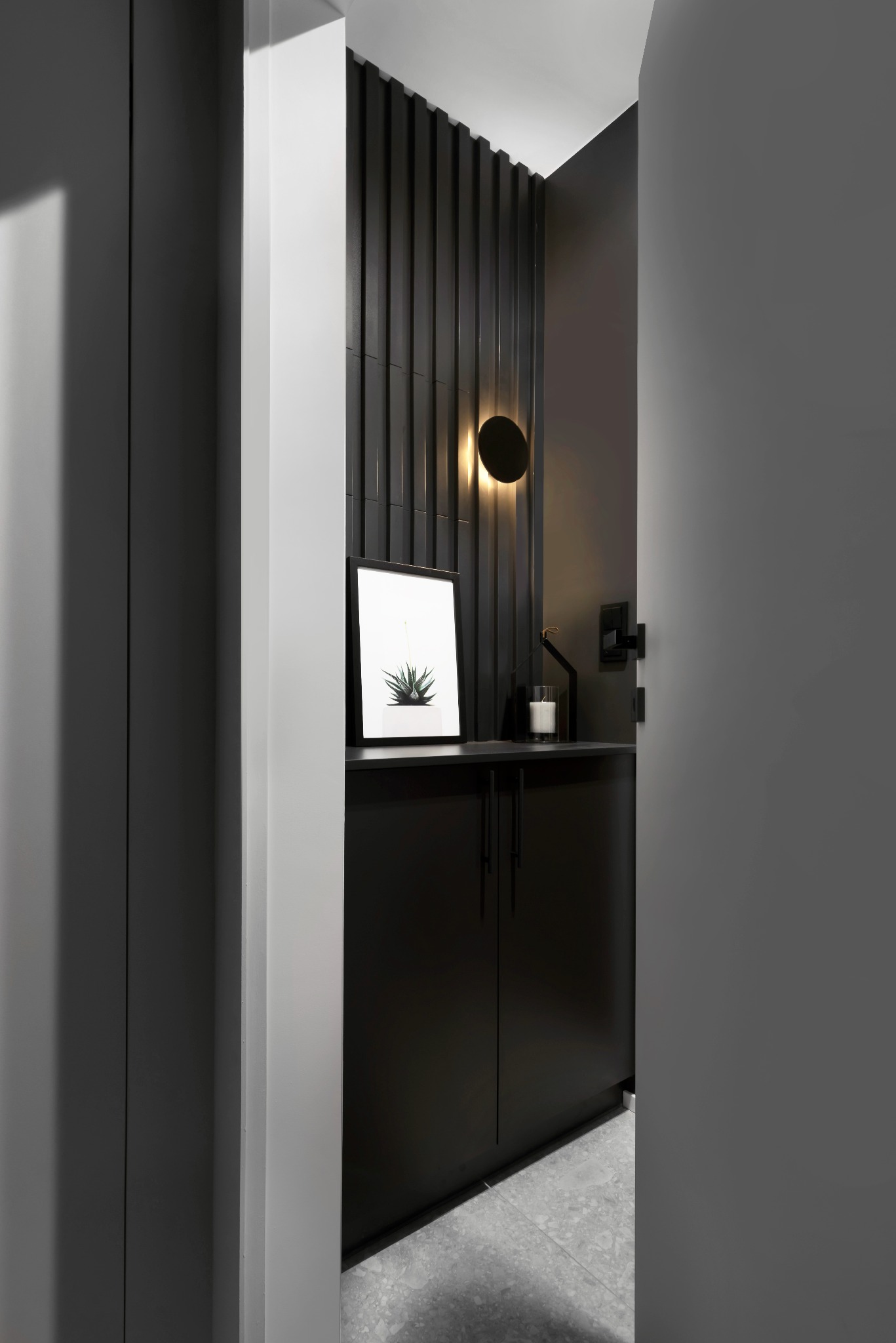 Nowoczesna, monochromatyczna łazienka z czarną szafką pod umywalką, okrągłym kinkietem i minimalistyczną grafiką z motywem roślinnym, widoczna przez częściowo otwarte drzwi.