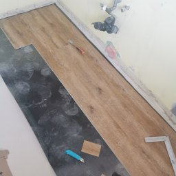 Rud bud - Fragment układania paneli podłogowych w pomieszczeniu, widoczne narzędzia: nożyk, młotek, kątownik, częściowo ułożone panele o wzorze drewna oraz ślady stóp na podkładzie.