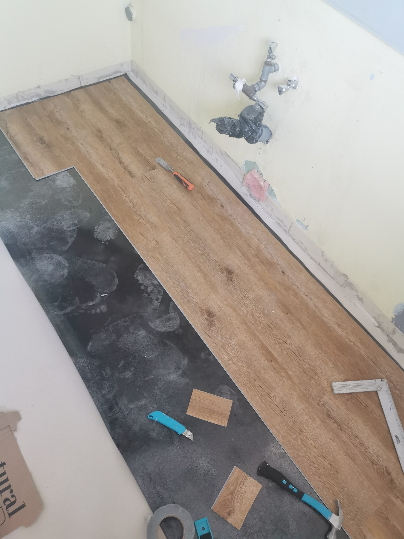 Fragment układania paneli podłogowych w pomieszczeniu, widoczne narzędzia: nożyk, młotek, kątownik, częściowo ułożone panele o wzorze drewna oraz ślady stóp na podkładzie.