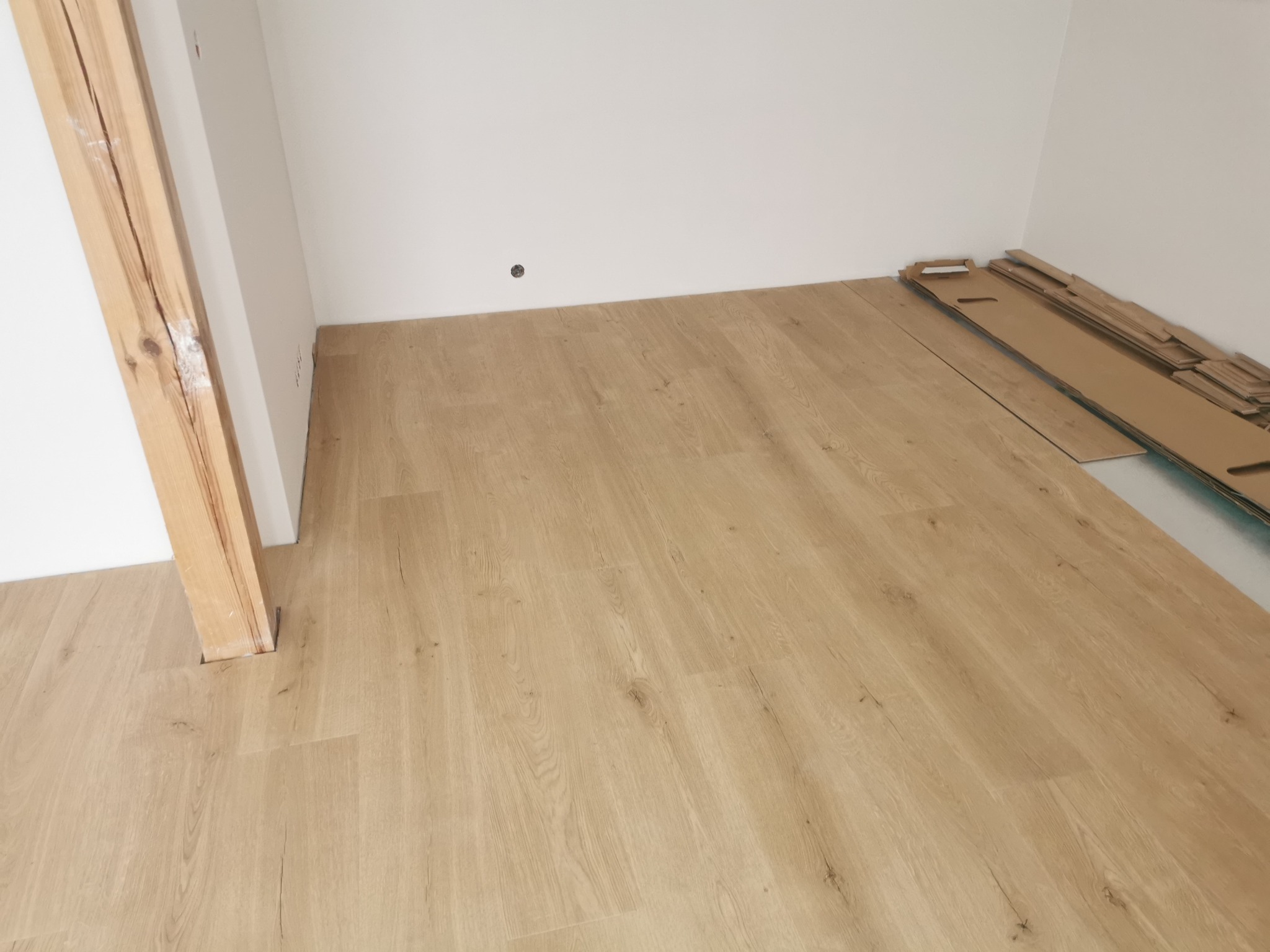 Fragment pokoju z jasnymi panelami podłogowymi ułożonymi wokół drewnianej futryny, z kartonami paneli w tle.