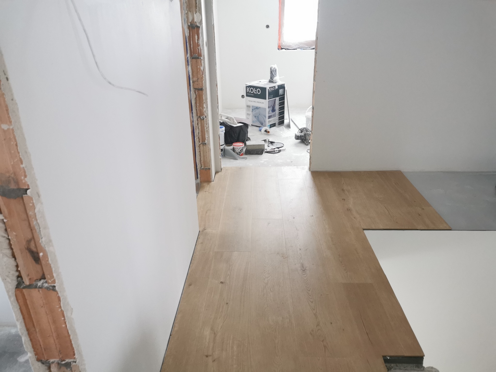 Montaż paneli podłogowych w trakcie, widoczne narzędzia i materiały budowlane w tle, surowy stan ścian i niedokończone wykończenie ościeżnicy.