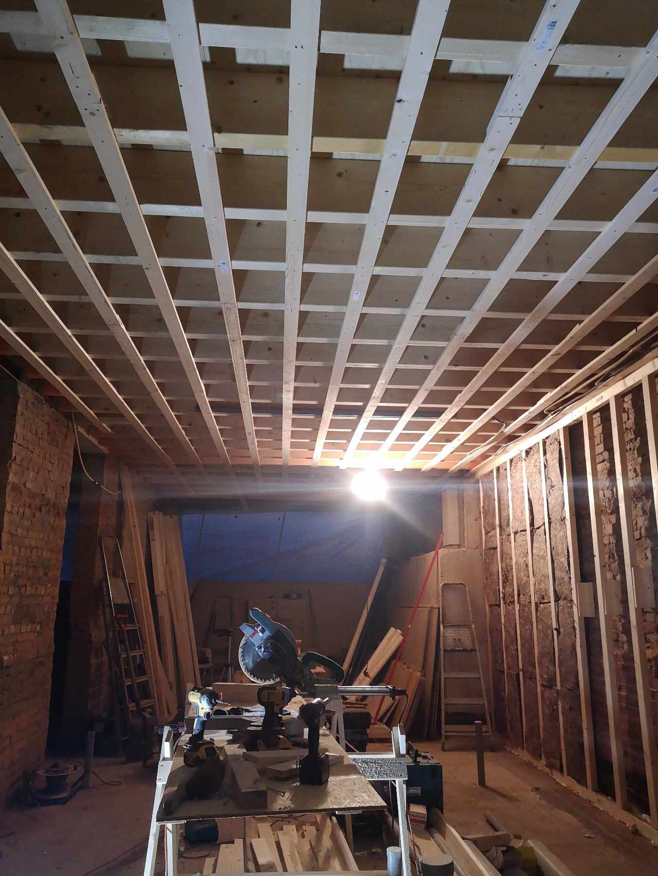 Konstrukcja drewniana sufitu podwieszanego w trakcie montażu, widoczne profile i płyty, w tle narzędzia i oświetlenie budowlane, surowe ściany z cegły i izolacji.