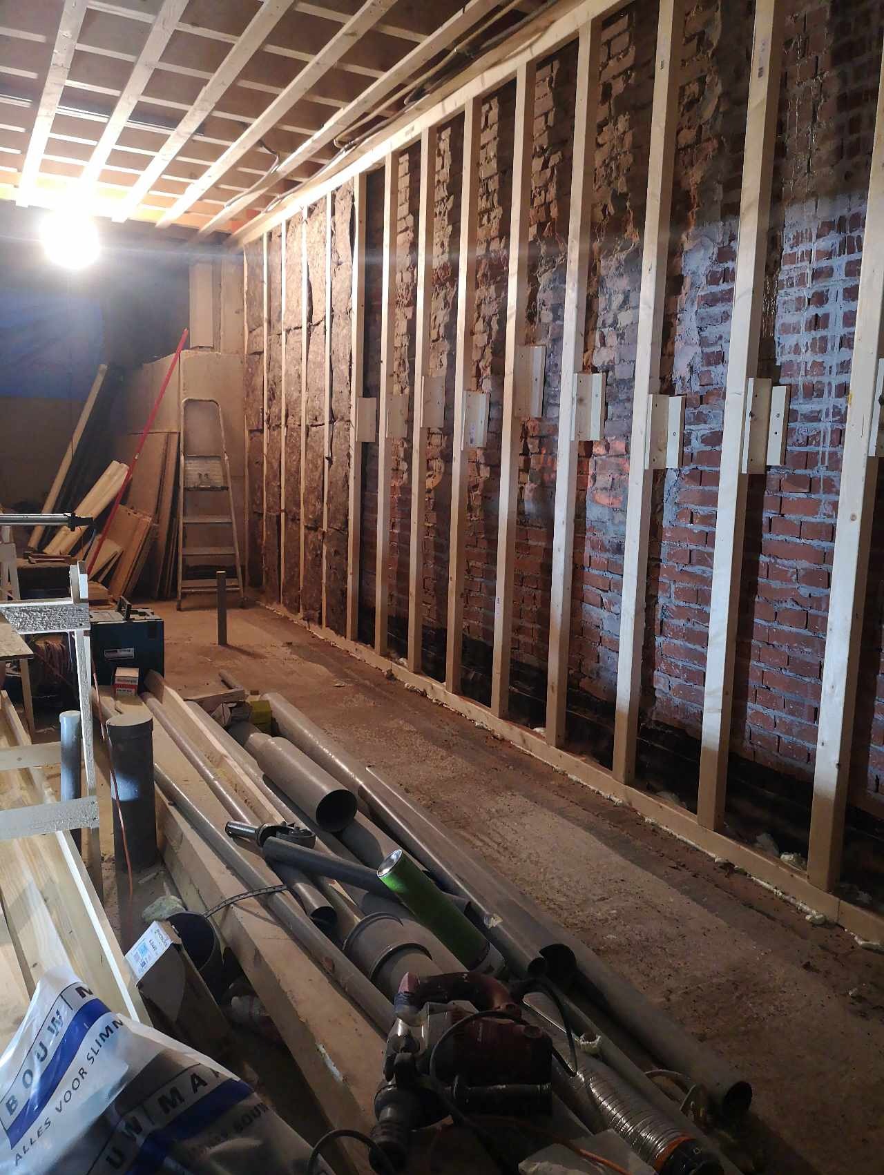 Wnętrze w trakcie prac adaptacyjnych: drewniana konstrukcja szkieletowa przy ceglanej ścianie, widoczne rury kanalizacyjne i inne materiały budowlane, surowy stan wykończenia.