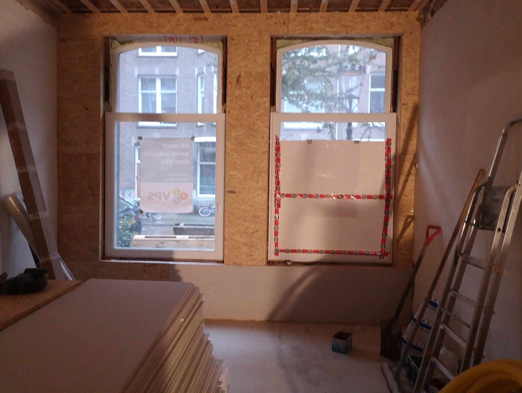 Przygotowanie do wykończenia wnętrza: dwa okna z osadzonymi ościeżnicami w surowej obudowie z płyt OSB, jedno okno zabezpieczone folią z taśmą, w tle sterta płyt gipsowych, drabina i narzędzia.