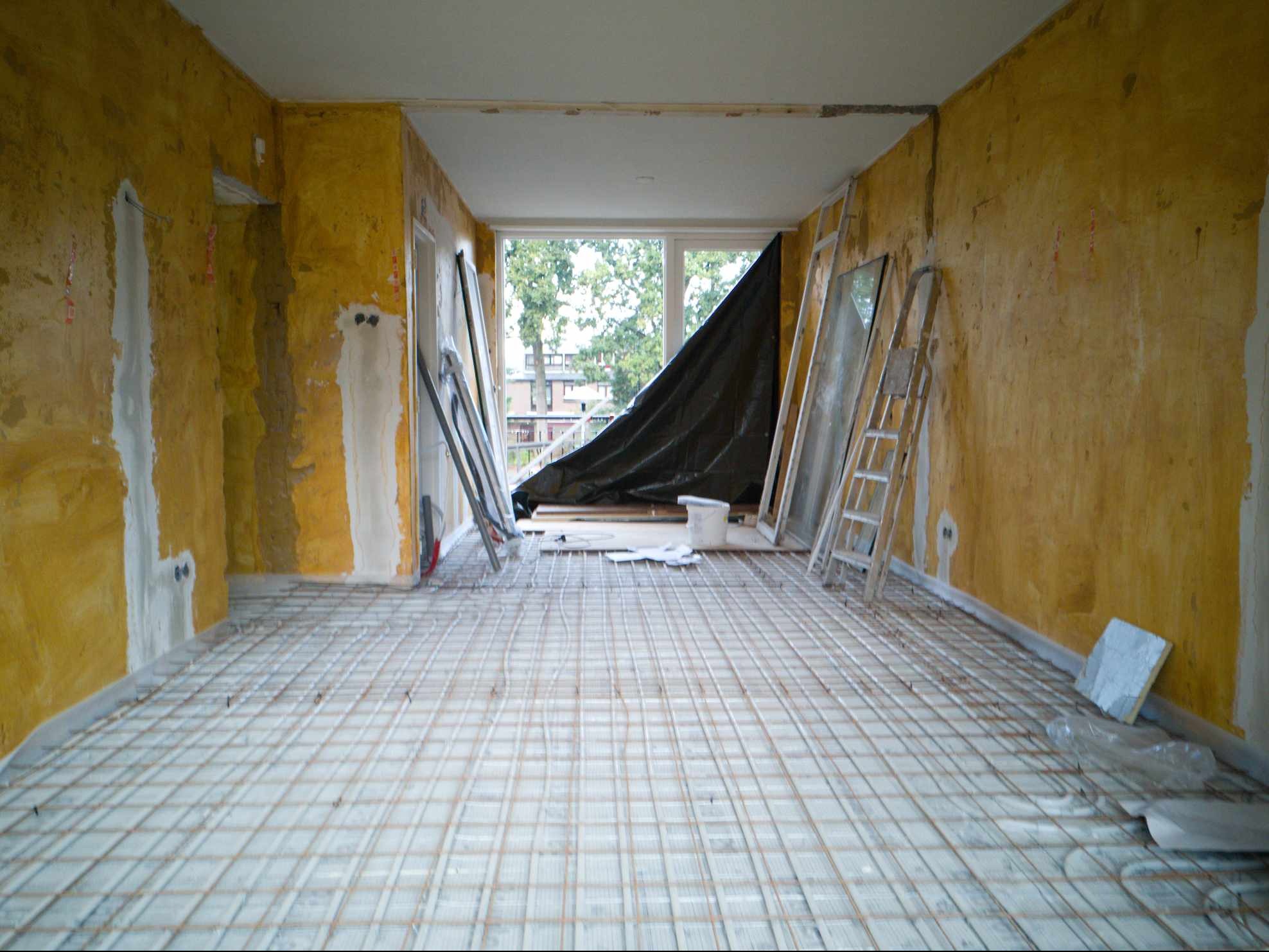 Pokój w trakcie remontu: przygotowane ściany do malowania z widocznymi śladami po szpachlowaniu, okno zabezpieczone folią, na podłodze siatka z przewodami ogrzewania podłogowego.
