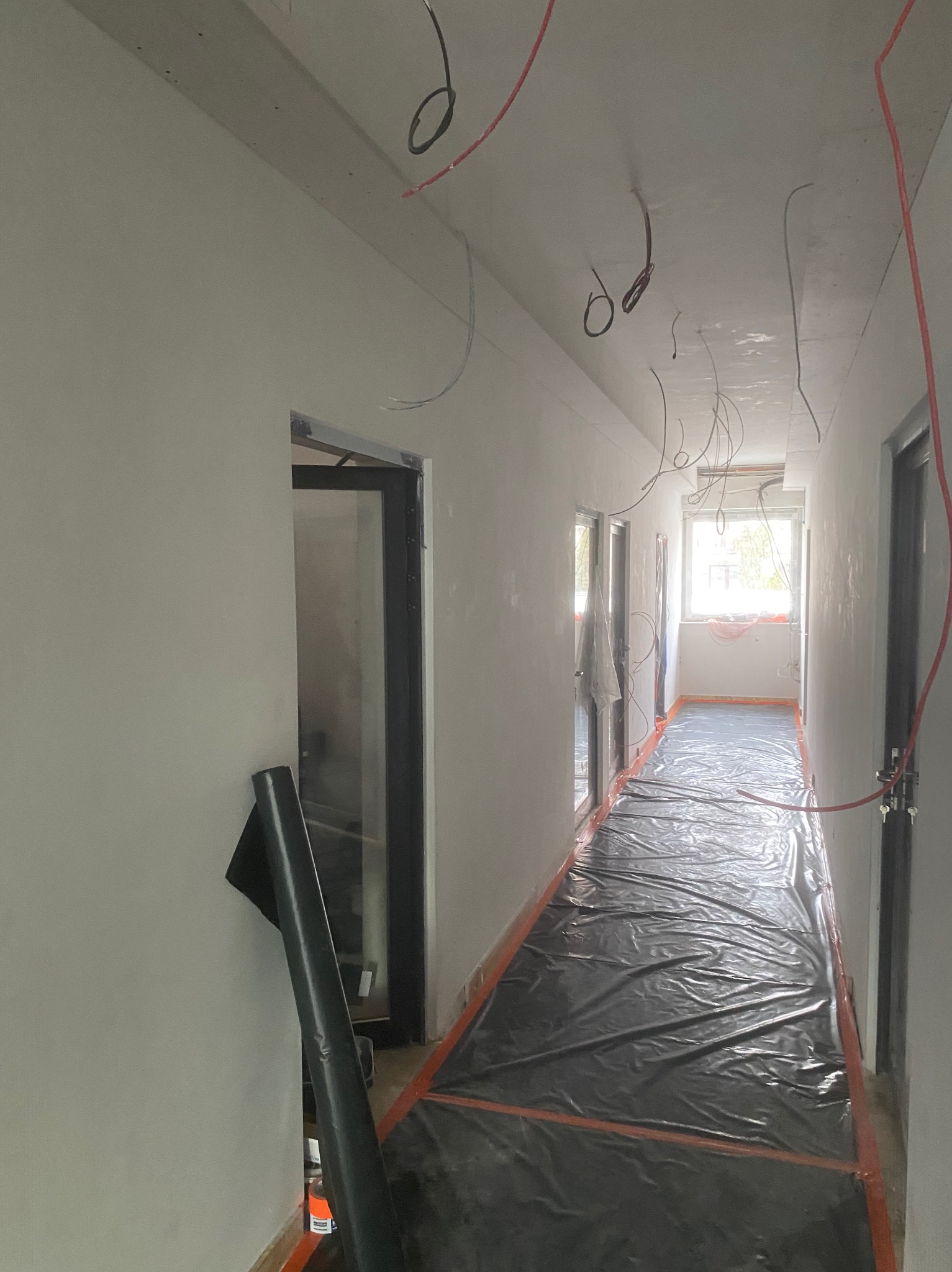 Korytarz w trakcie prac przygotowawczych: ściany po szpachlowaniu, zabezpieczona folią podłoga, wystające przewody elektryczne z sufitu, czarne drzwi z ościeżnicami.