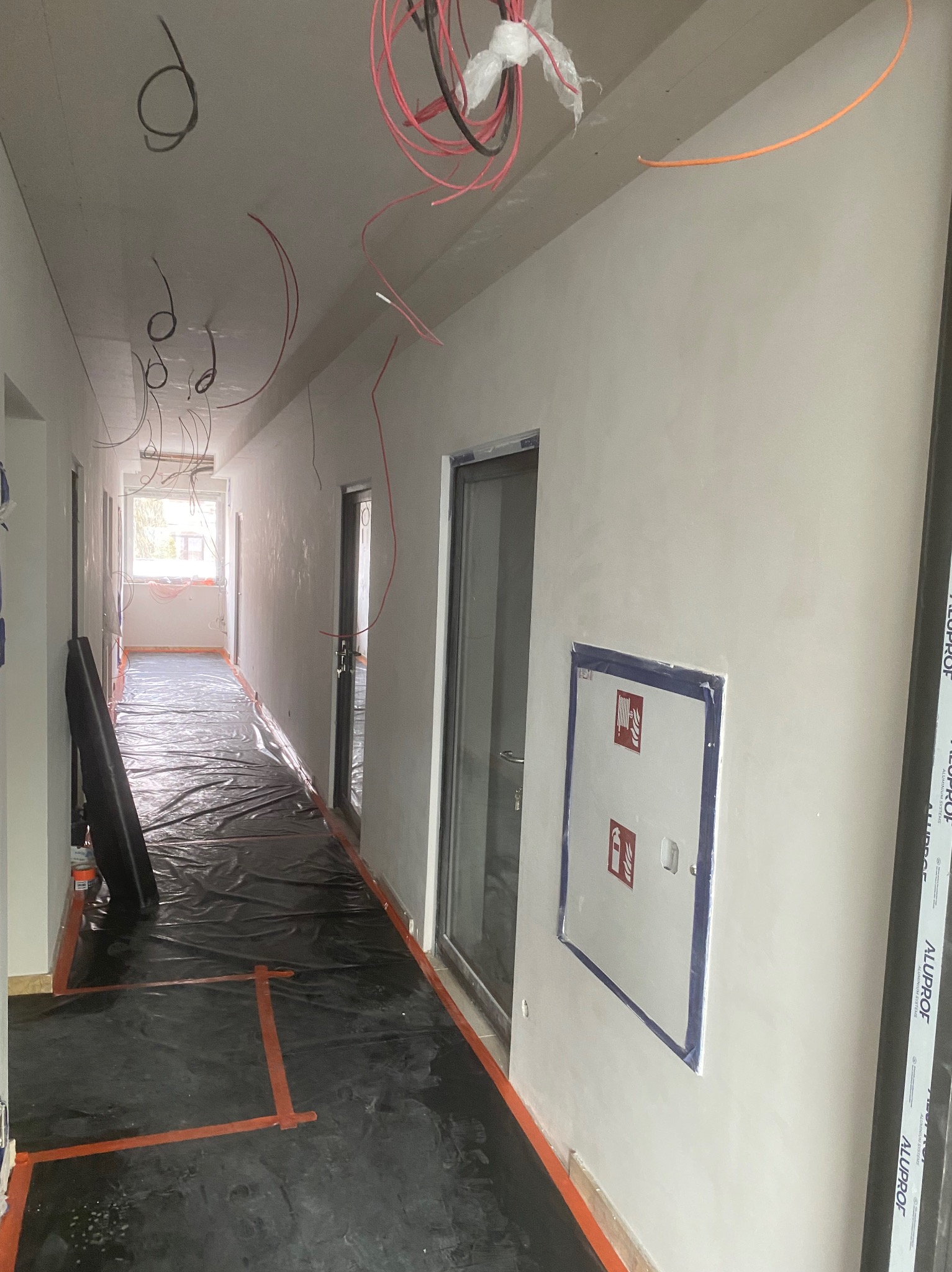 Korytarz w trakcie prac wykończeniowych: zabezpieczona folią podłoga z taśmą, świeżo wyszpachlowane ściany z widoczną skrzynką hydrantową, zwisające kable instalacji elektrycznej.