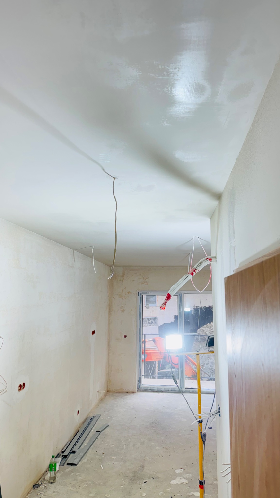 Wnętrze w trakcie prac wykończeniowych: ściany po szpachlowaniu, widoczne przewody elektryczne, listwy przypodłogowe i oświetlenie budowlane. Etap przygotowania do malowania.