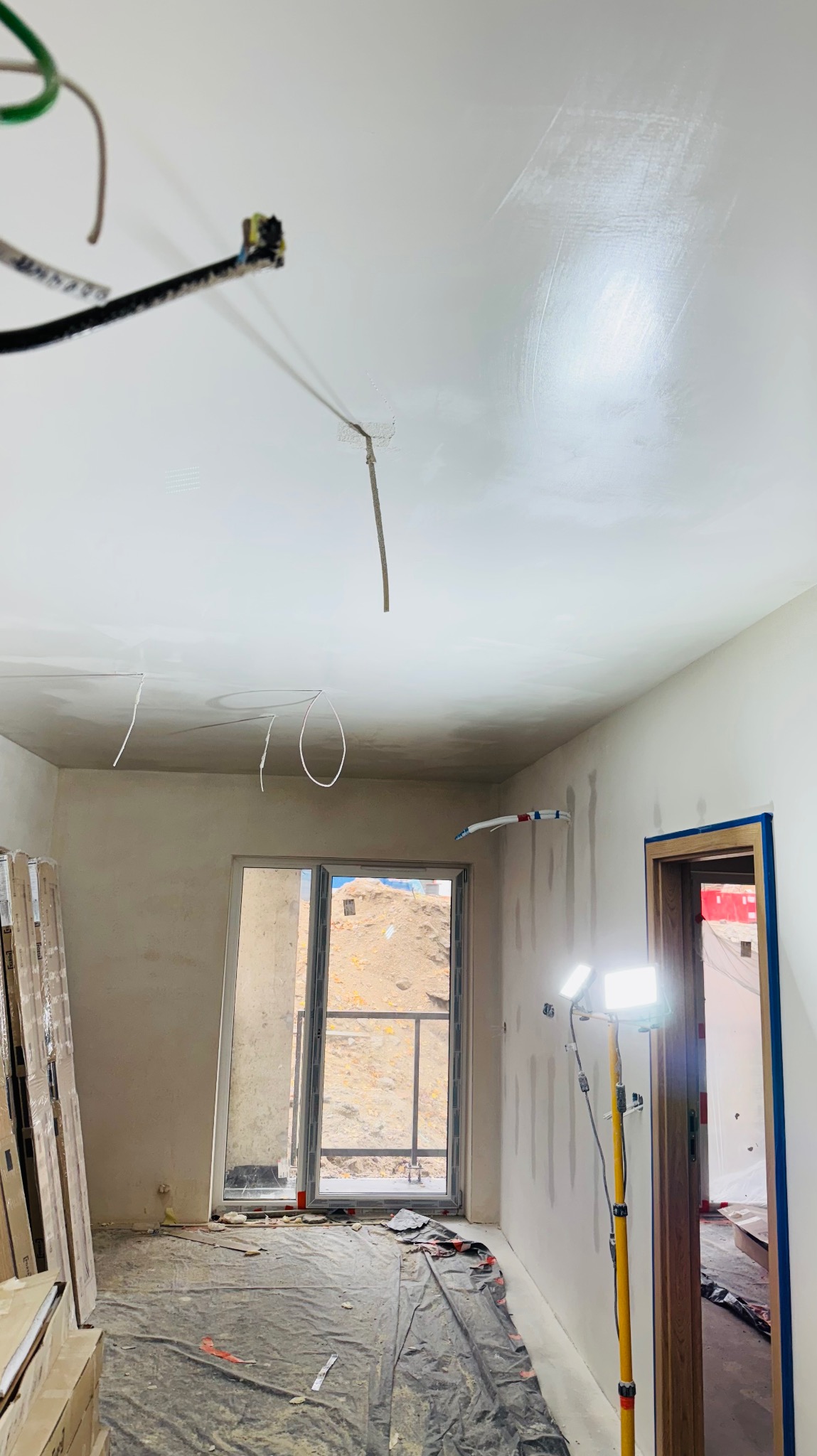 Wnętrze w trakcie remontu: gładzie na ścianach i suficie, widoczne przewody elektryczne, okno z widokiem na zewnątrz, podłoga zabezpieczona folią, lampa budowlana.