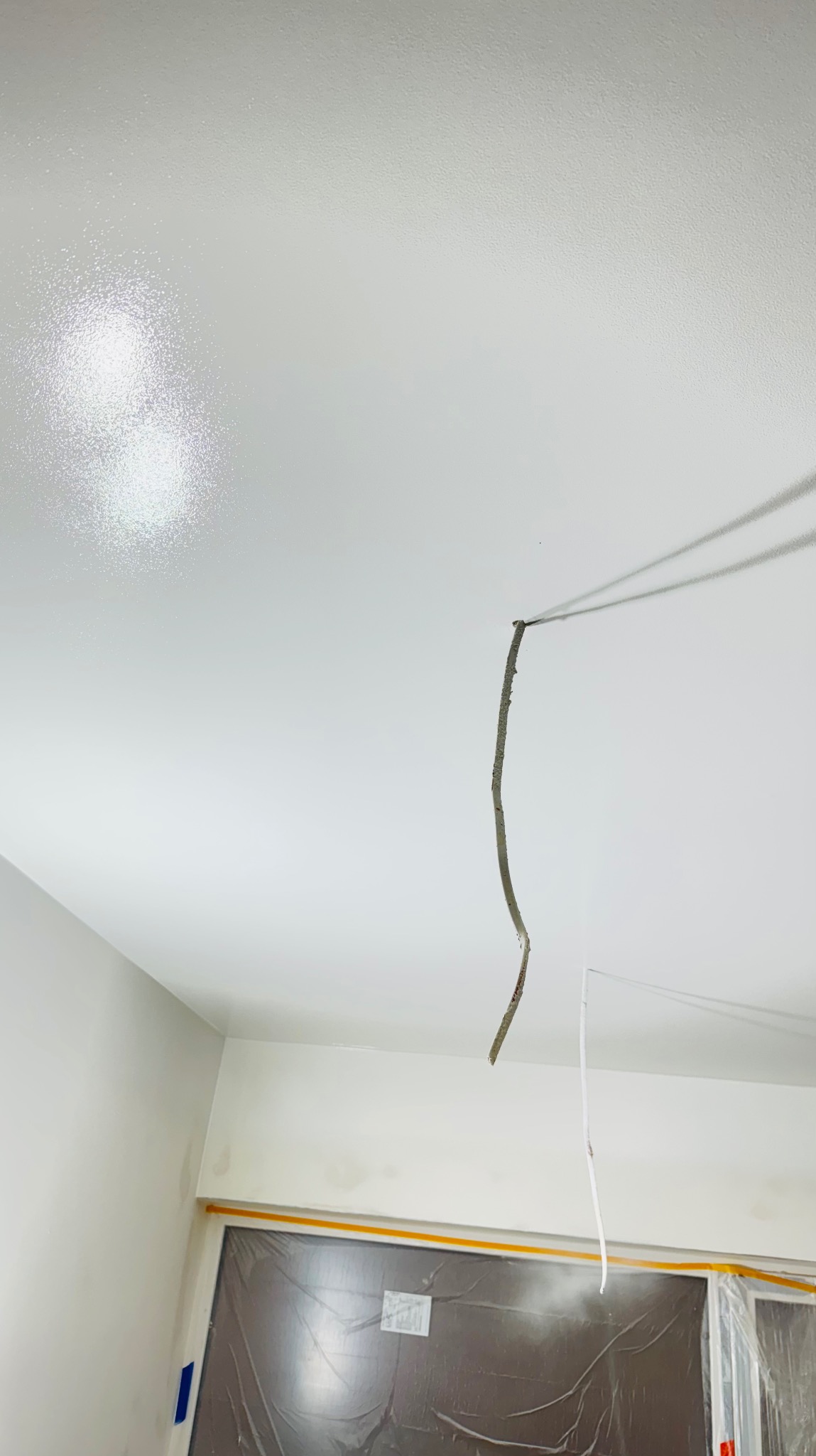 Biały sufit po szpachlowaniu z wystającym przewodem elektrycznym i zabezpieczonym oknem folią malarską. Widoczne ślady po łataniu.