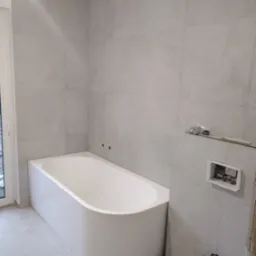 Wanna z obudową i szare płytki w trakcie montażu w łazience w Grójcu, widoczne niezamaskowane otwory na rury i przygotowane miejsce na panel sterowania.