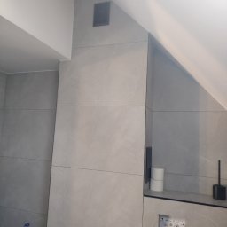 ART-BUD Usługi ogólnobudowlane - Ściana łazienki w szarych płytkach imitujących kamień, zabudowa stelaża WC z widocznymi przyciskami spłukiwania, półka z zapasem papieru toaletowego i szczotką do WC, wentylacja w suficie pod skosem.