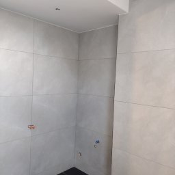 ART-BUD Usługi ogólnobudowlane - Wnętrze łazienki w trakcie wykańczania, widoczne szare płytki imitujące marmur na ścianach, czarna podłoga, wystające przyłącza wodne i elektryczne.