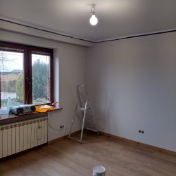 ART-BUD Usługi ogólnobudowlane - Pokój w trakcie przygotowania do malowania: zabezpieczone taśmą krawędzie sufitu, drabina oparta o ścianę, wiadro z farbą na podłodze, grzejnik pod oknem z radiem i kuwetą malarską.