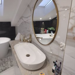 ART-BUD Usługi ogólnobudowlane - Umywalka nablatowa o owalnym kształcie z baterią w kolorze złotym, zamontowana na blacie z jasnego kamienia w łazience z marmurowymi płytkami i owalnym lustrem w złotej ramie, w tle widoczna wanna.