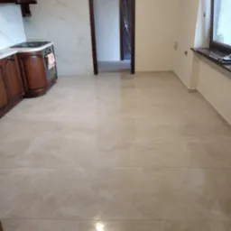 Wykończona podłoga z płytek imitujących marmur w jasnej tonacji, widok na otwarte przejście do innego pomieszczenia i fragment zabudowy kuchennej z drewnianymi szafkami.