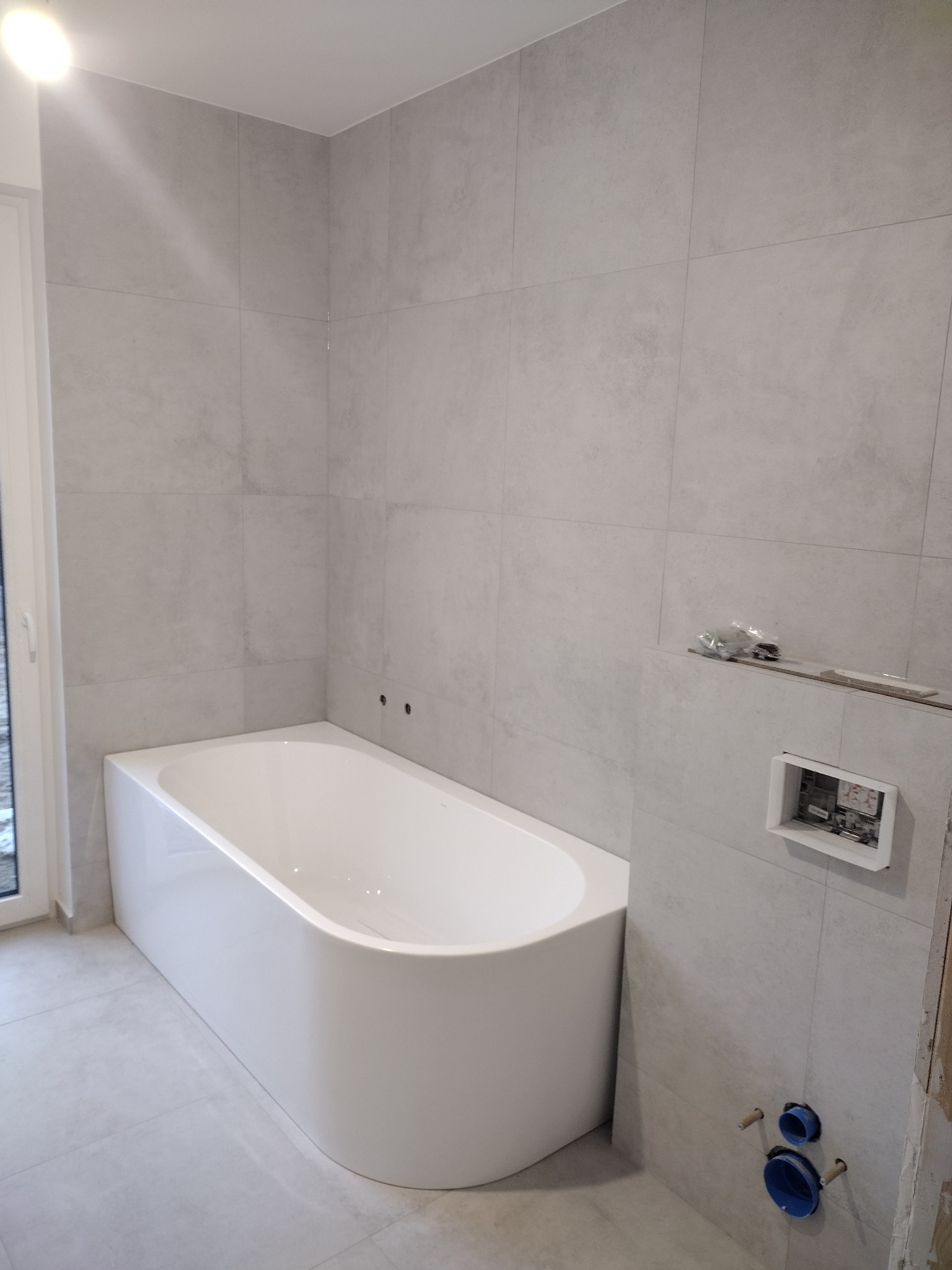 Wanna z obudową i szare płytki w trakcie montażu w łazience w Grójcu, widoczne niezamaskowane otwory na rury i przygotowane miejsce na panel sterowania.