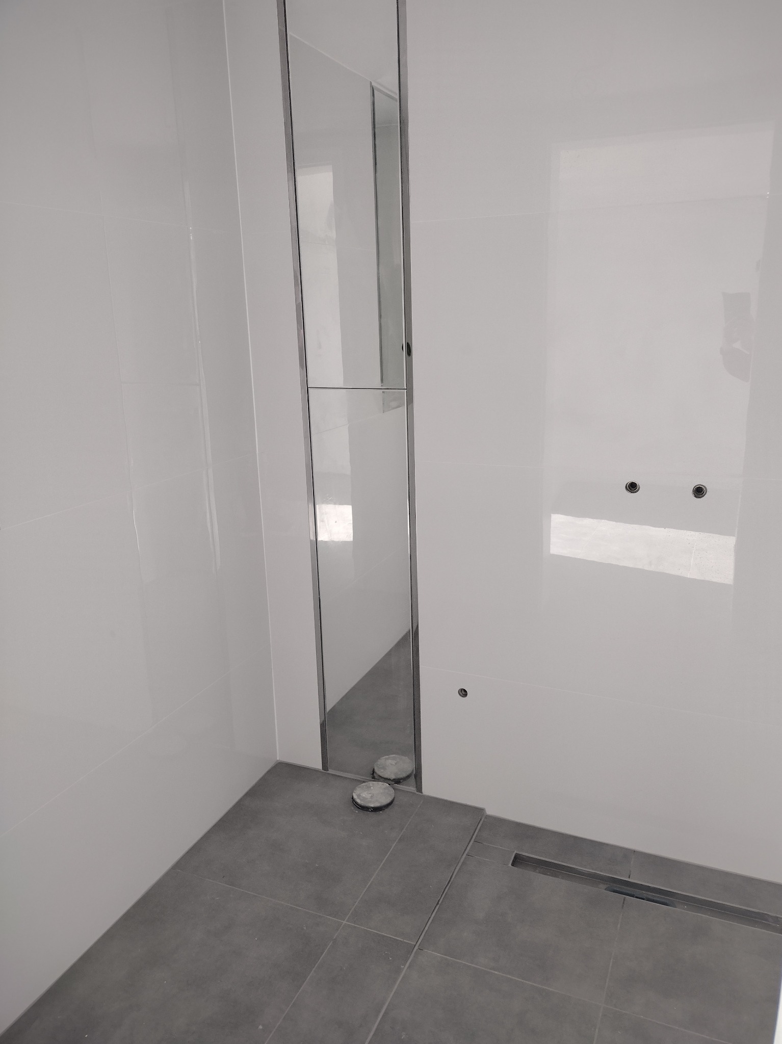 Nowoczesna łazienka z białymi błyszczącymi płytkami ściennymi, szarą podłogą z odpływem liniowym i szklaną kabiną prysznicową z chromowanymi profilami.