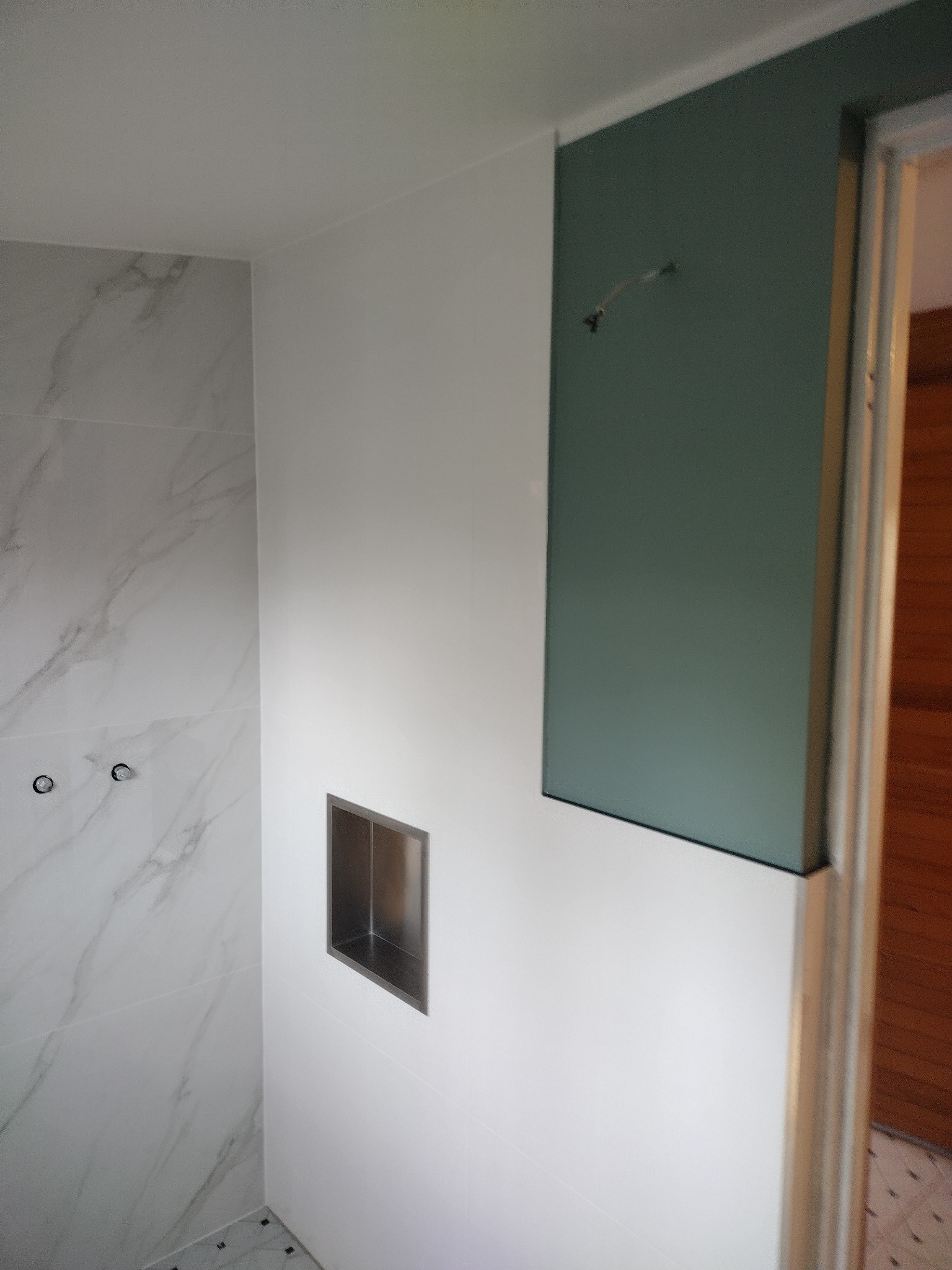 Wnętrze łazienki w trakcie prac wykończeniowych: białe marmurowe płytki na ścianie, wnęka w ścianie z metalowym obramowaniem, fragment ściany pomalowany na ciemnozielono, widoczne przewody...