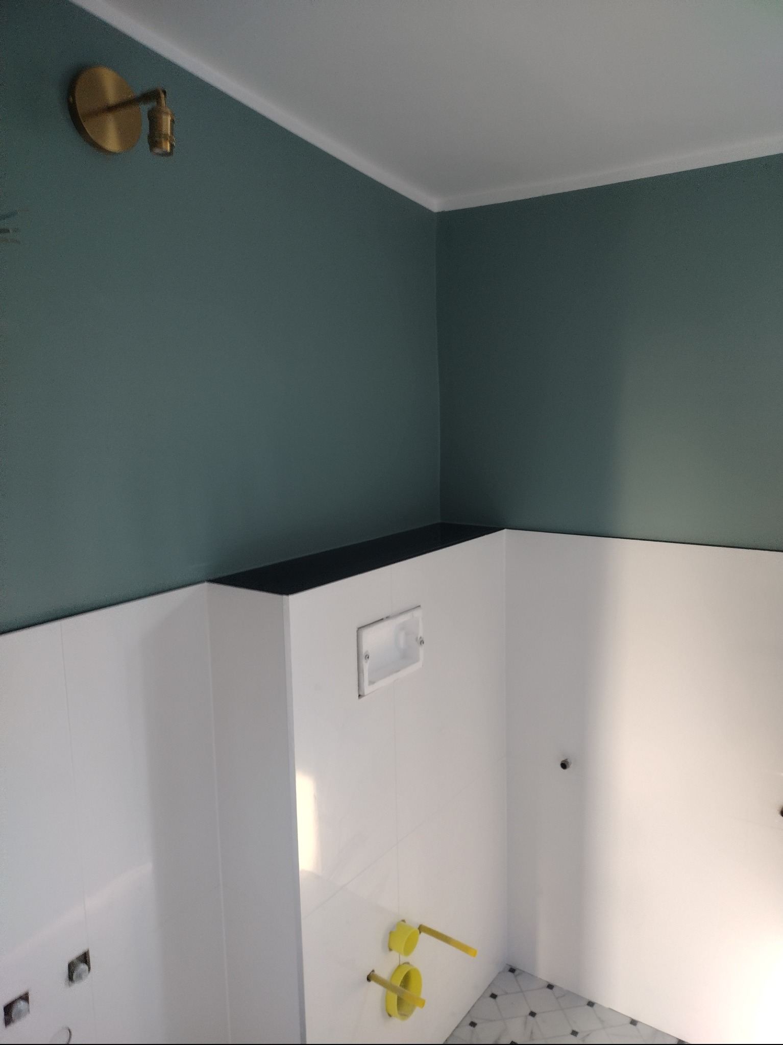 Świeżo wyremontowana łazienka z białymi płytkami ściennymi, zieloną górną częścią ścian, złotą lampą ścienną i czarnym blatem, widoczny stelaż podtynkowy WC oraz fragment podłogi z szaro-białą...