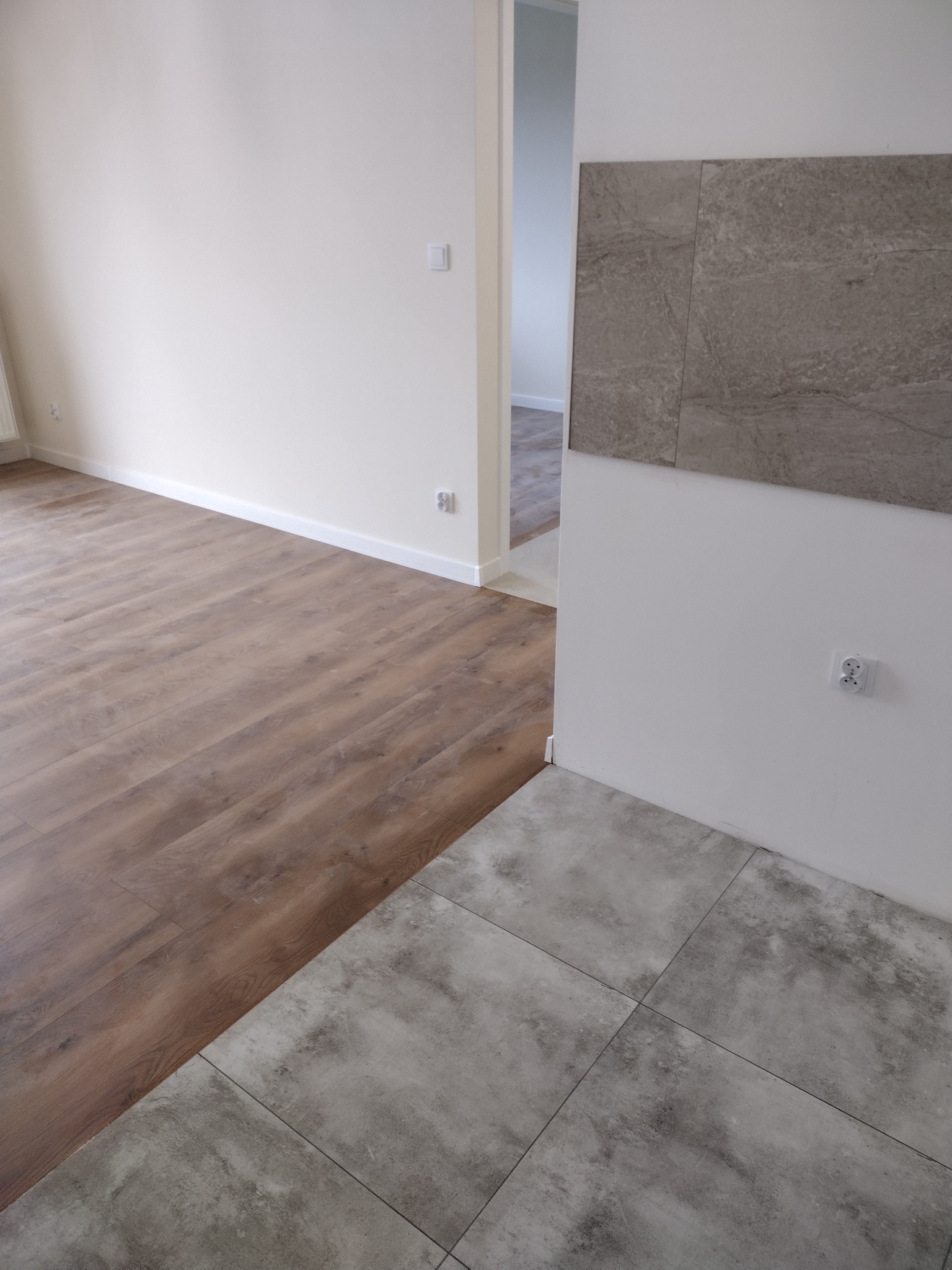 Połączenie paneli podłogowych imitujących drewno z szarą, matową płytką podłogową, widoczne gniazdka elektryczne i fragment ceramicznej okładziny ściennej w odcieniach szarości, jasne ściany.