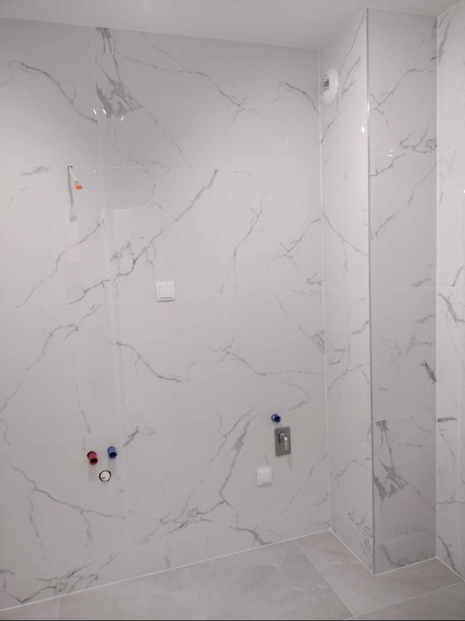 Wnętrze łazienki w trakcie prac wykończeniowych, widoczne marmurowe płytki na ścianach, szare płytki podłogowe, przygotowane przyłącza wodne, wentylator w suficie i włącznik światła.