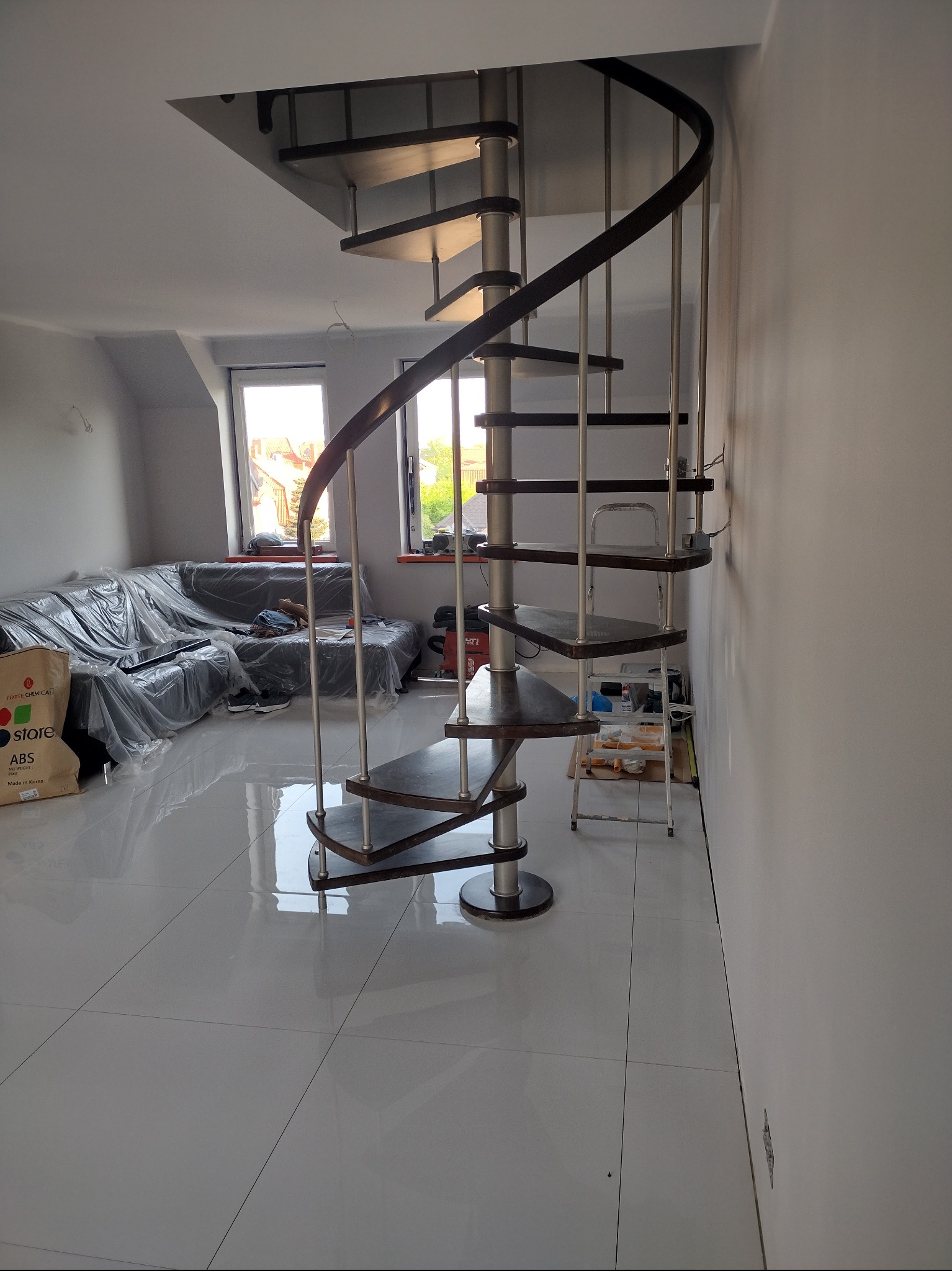 Spiralne schody z drewnianymi stopniami i metalową konstrukcją w jasnym pomieszczeniu z błyszczącą podłogą, kanapa przykryta folią ochronną, widoczne okna i elementy wyposażenia.