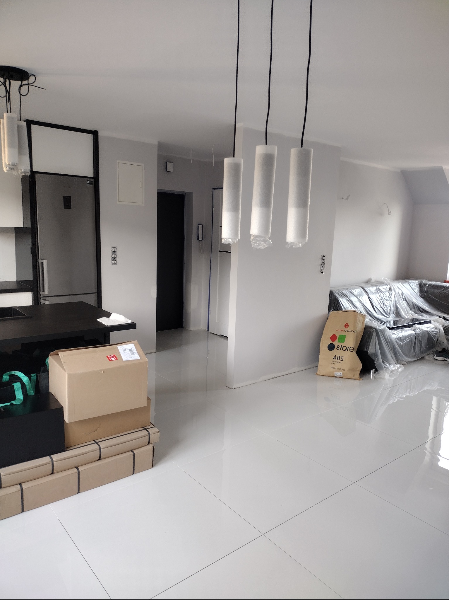 Wnętrze w trakcie prac wykończeniowych: białe, błyszczące płytki podłogowe, zabezpieczone meble folią, kartony z materiałami budowlanymi i lampy wiszące.