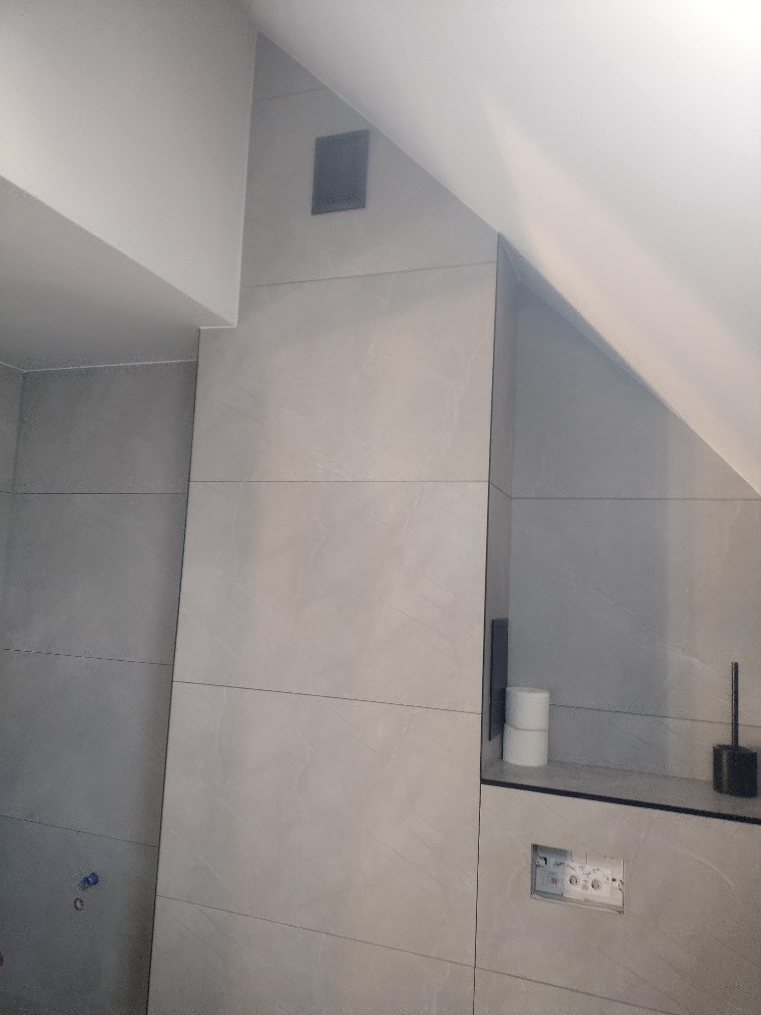 Ściana łazienki w szarych płytkach imitujących kamień, zabudowa stelaża WC z widocznymi przyciskami spłukiwania, półka z zapasem papieru toaletowego i szczotką do WC, wentylacja w suficie pod skosem.