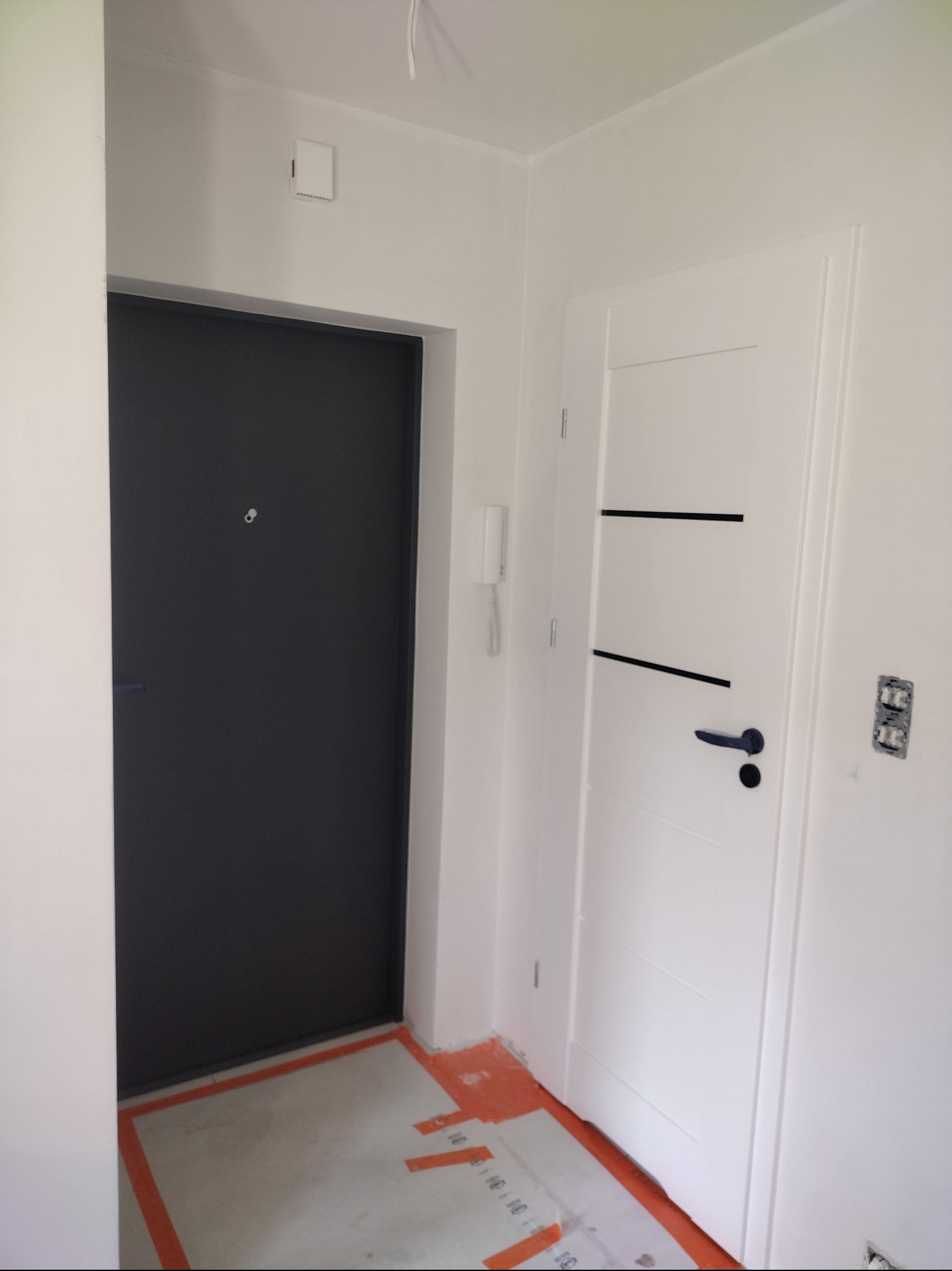 Wnętrze mieszkania w trakcie prac wykończeniowych: widok na zamontowane drzwi wejściowe w kolorze antracytowym oraz białe drzwi wewnętrzne z czarnymi poziomymi wstawkami, na podłodze widoczne...