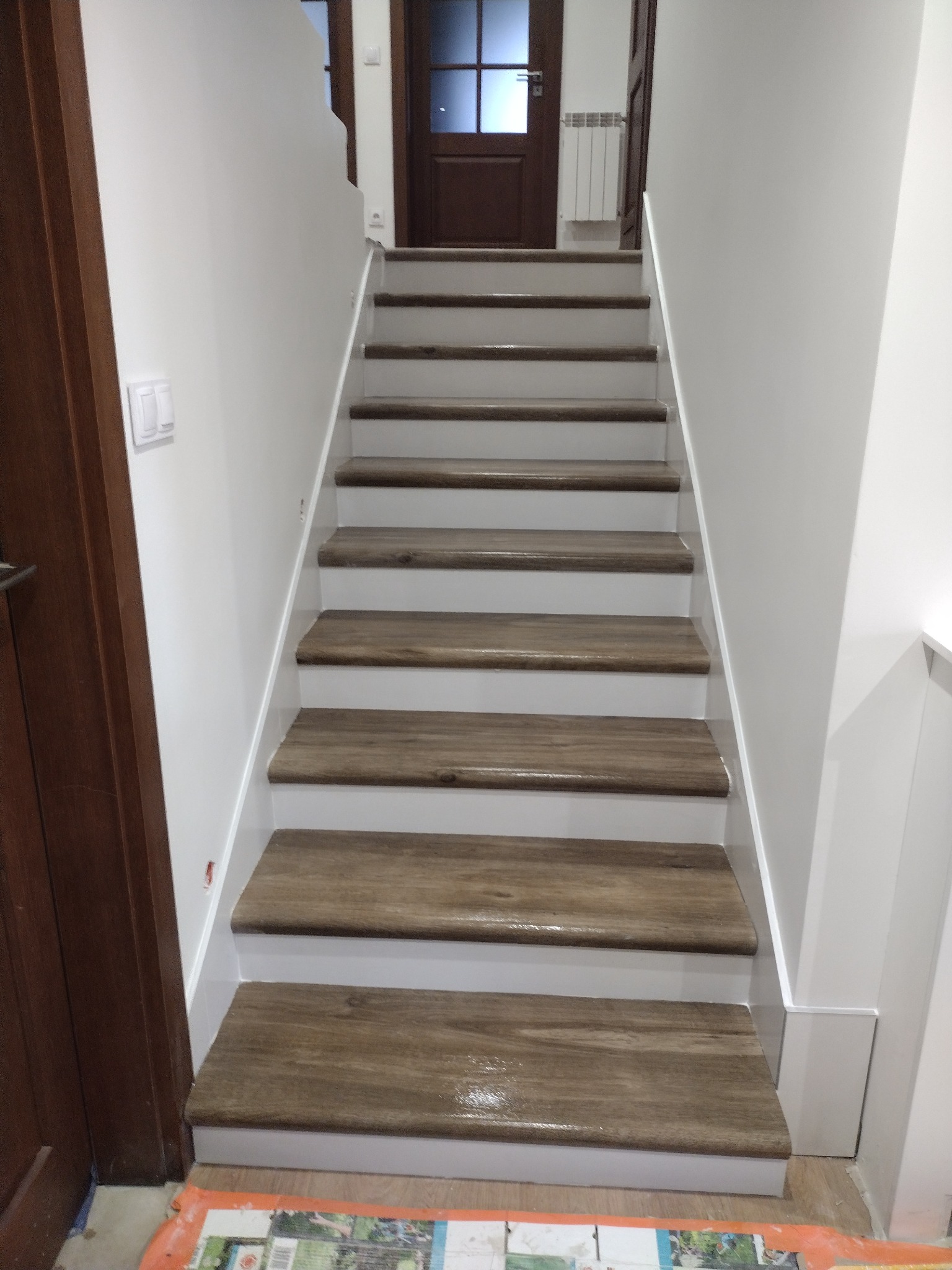 Schody z drewnianymi stopniami i białymi podstopnicami w trakcie prac wykończeniowych, zabezpieczone folią malarską na dole, widoczny fragment drzwi i grzejnika.