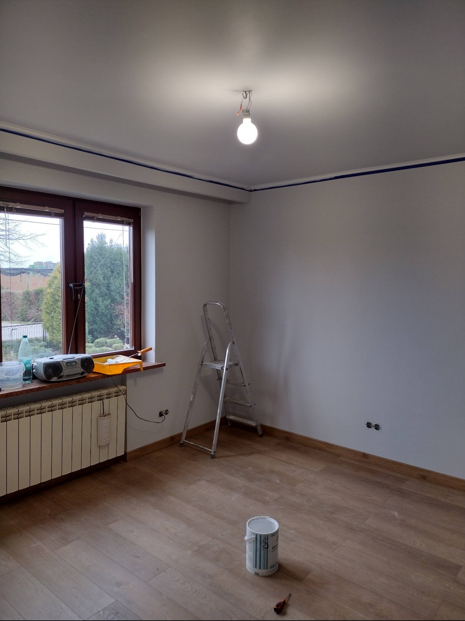 Pokój w trakcie przygotowania do malowania: zabezpieczone taśmą krawędzie sufitu, drabina oparta o ścianę, wiadro z farbą na podłodze, grzejnik pod oknem z radiem i kuwetą malarską.