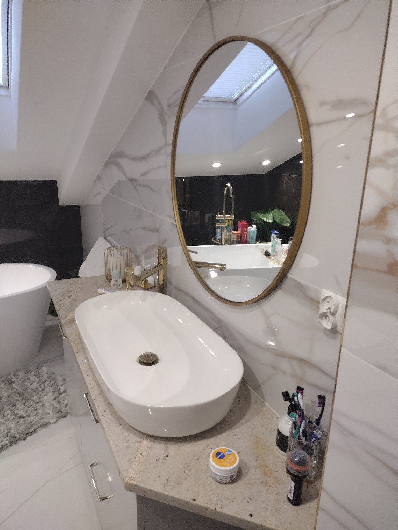 Umywalka nablatowa o owalnym kształcie z baterią w kolorze złotym, zamontowana na blacie z jasnego kamienia w łazience z marmurowymi płytkami i owalnym lustrem w złotej ramie, w tle widoczna wanna.