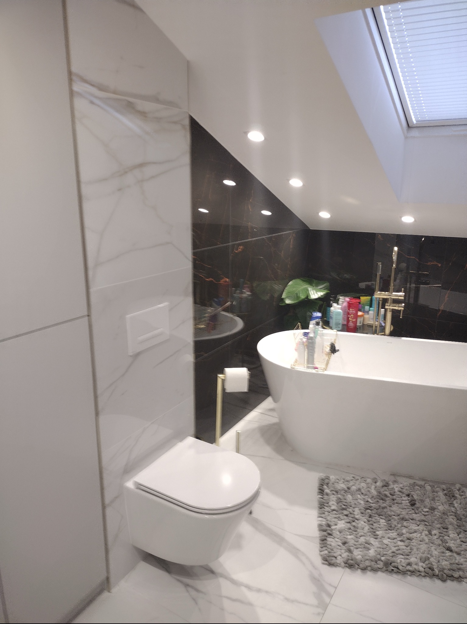 Nowoczesna łazienka z czarno-białymi marmurowymi płytkami, wiszącą toaletą, wolnostojącą wanną i oknem dachowym.